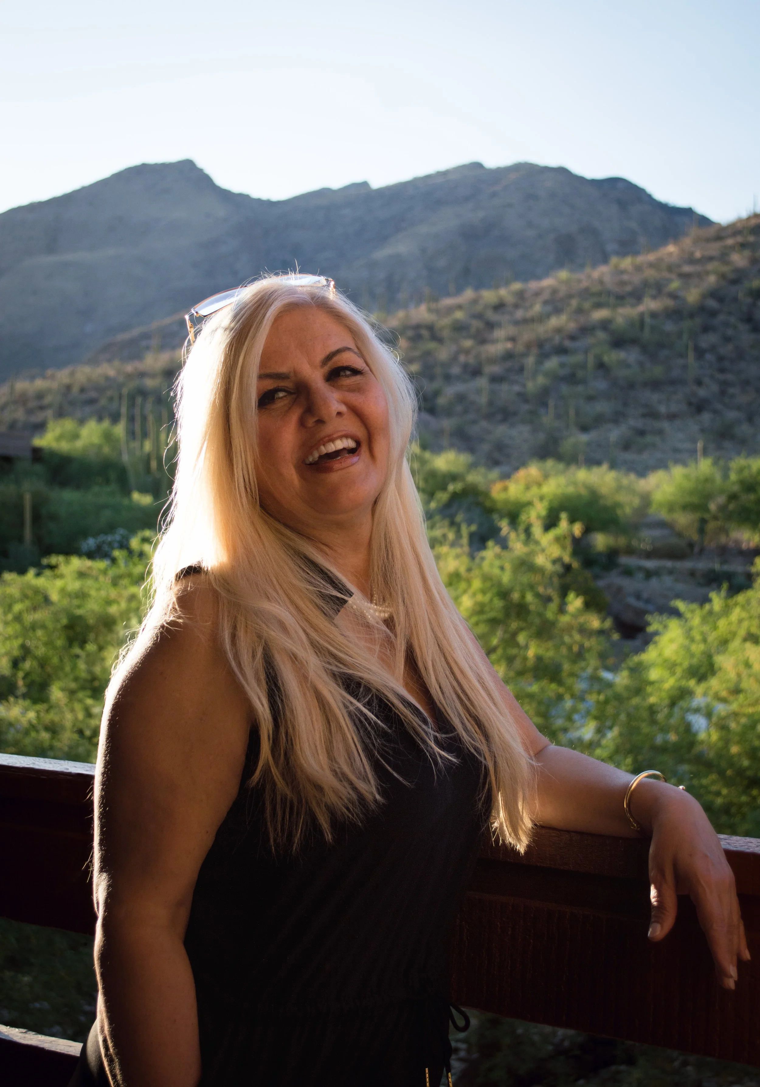 A woman with long blonde hair smiling and leaning on a wooden railing outdoors. Mountain landscape with green trees and bushes in the background, sunny sky.