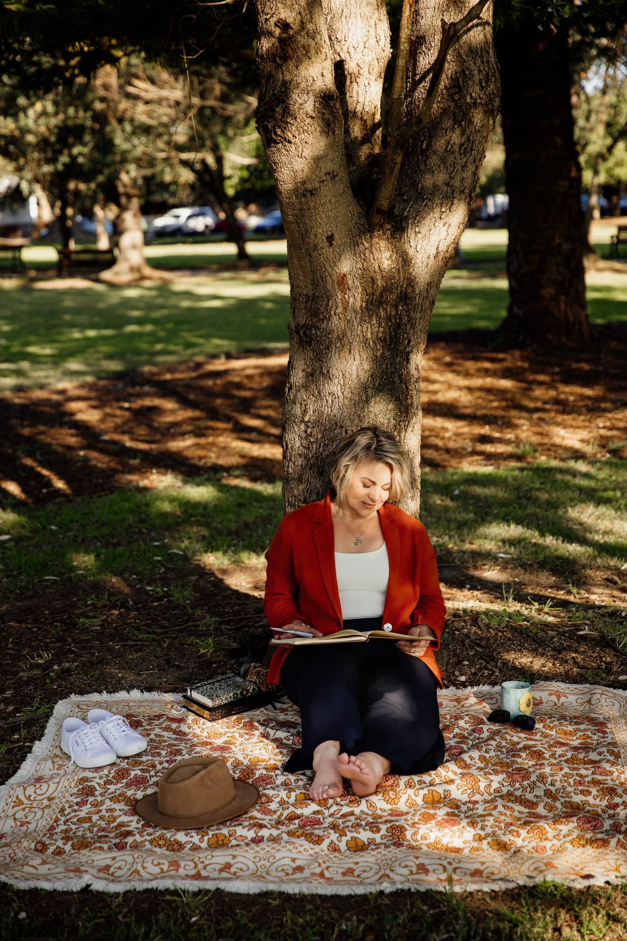 A woman sits on a patterned outdoor rug under a tree in a park, reading a book. She has short blonde hair, wears a white top, red blazer, and black pants. Nearby are white sneakers, a brown hat, a mug, sunglasses, and a closed book.