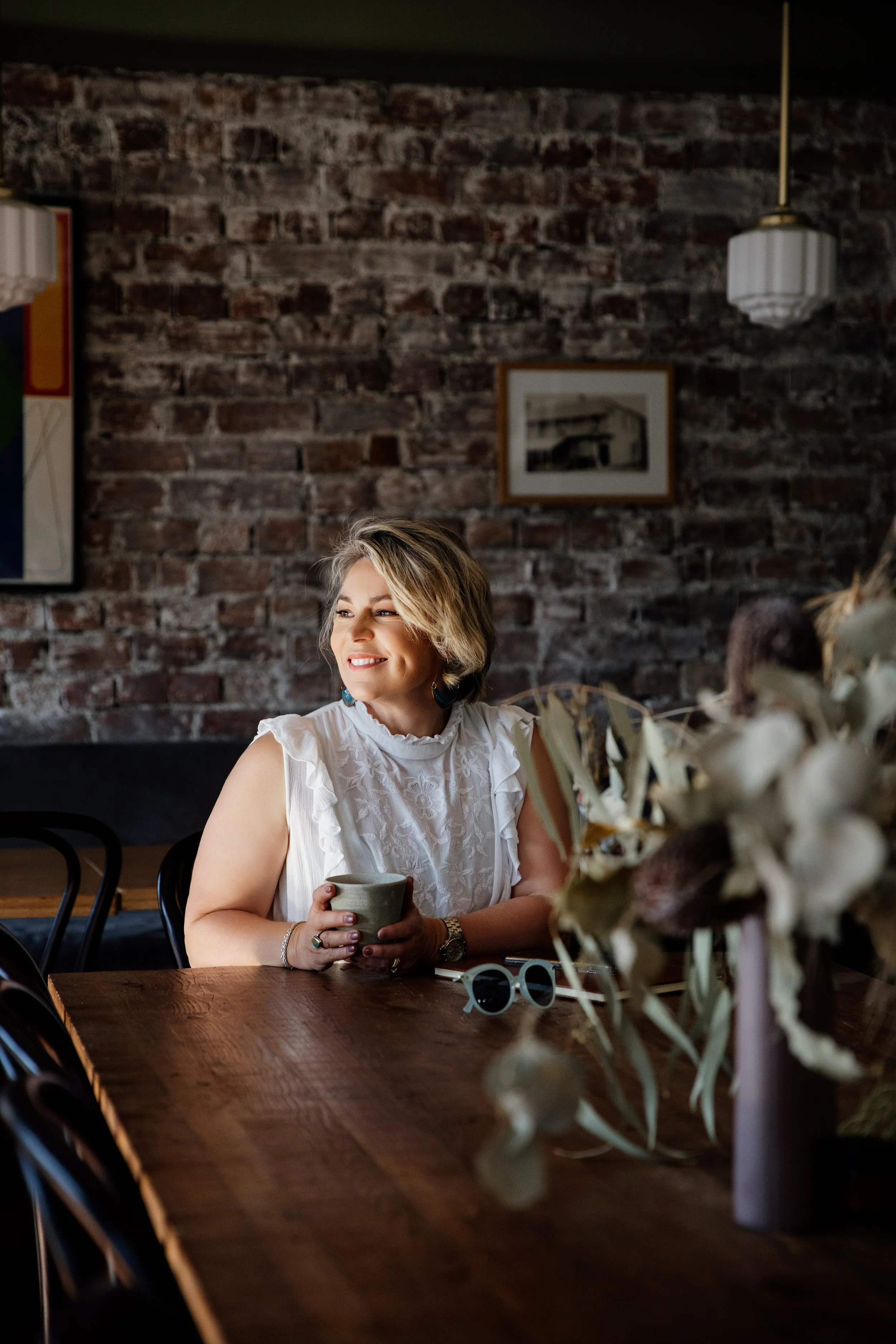 Woman wearing a white sleeveless top sitting at a wooden table in a cozy cafe with a brick wall background, holding a cup and smiling, with sunglasses and a floral arrangement on the table.