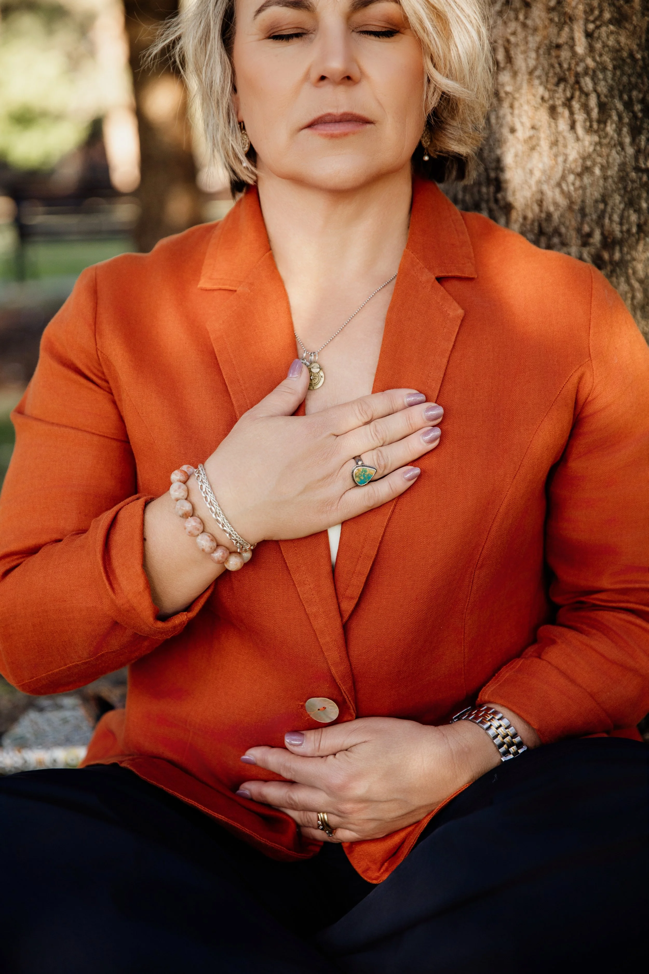 A woman with short blonde hair, wearing an orange blazer and jewelry, sitting outdoors with her eyes closed and hand on her chest and stomach.