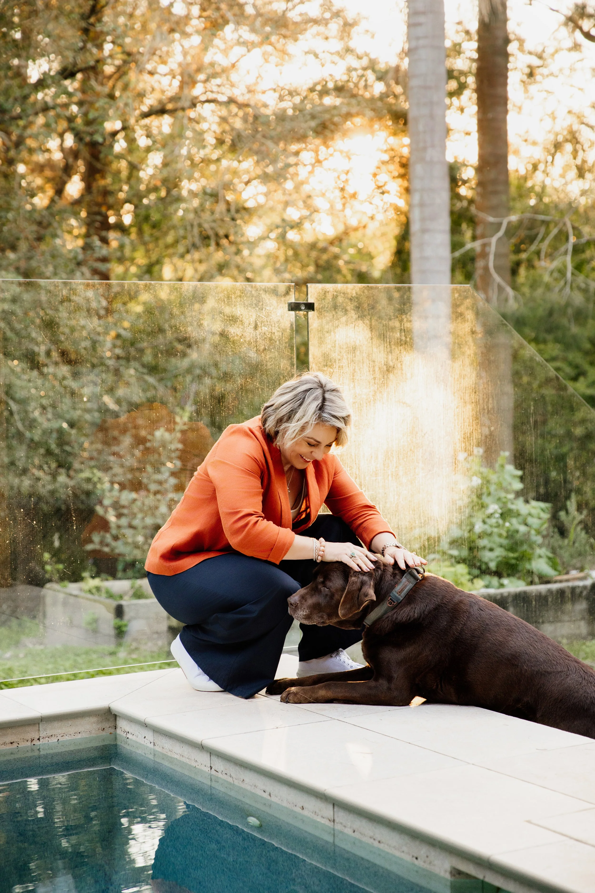A woman in an orange jacket and dark pants is kneeling by a large brown dog, petting it, on a poolside patio with plants and trees in the background during sunset.
