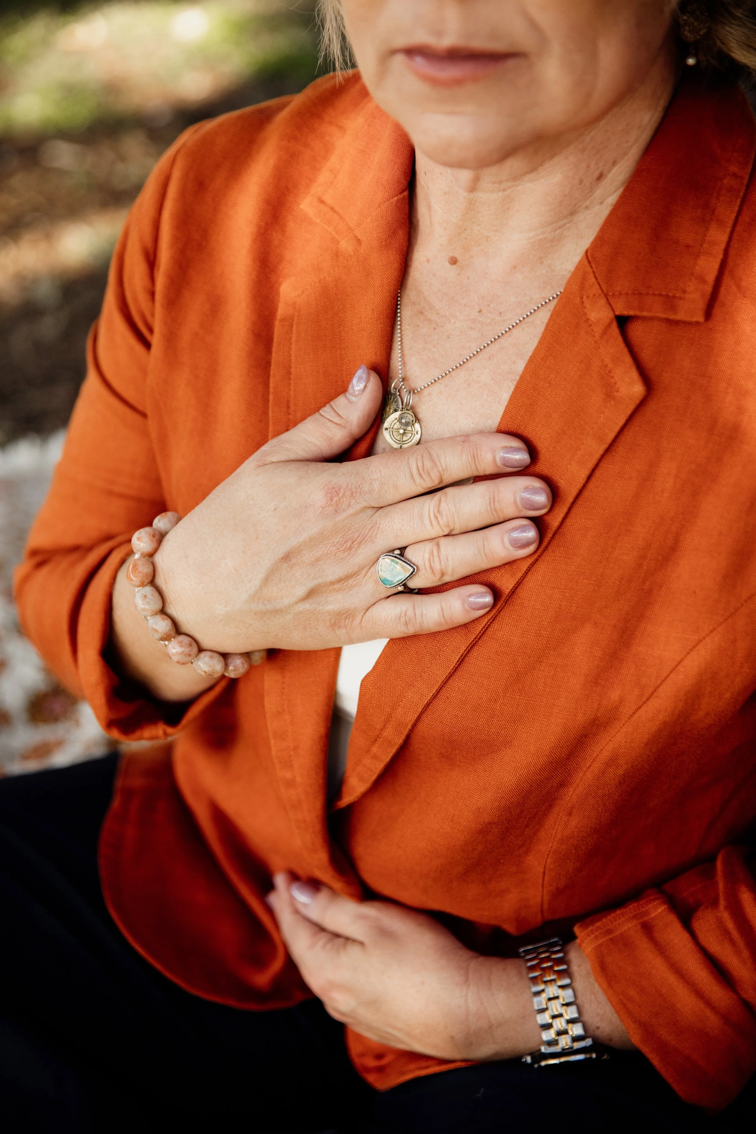 Close-up of a woman wearing an orange blazer, with her left hand on her chest showing jewelry including a necklace, ring, and bracelet. She is sitting outdoors.