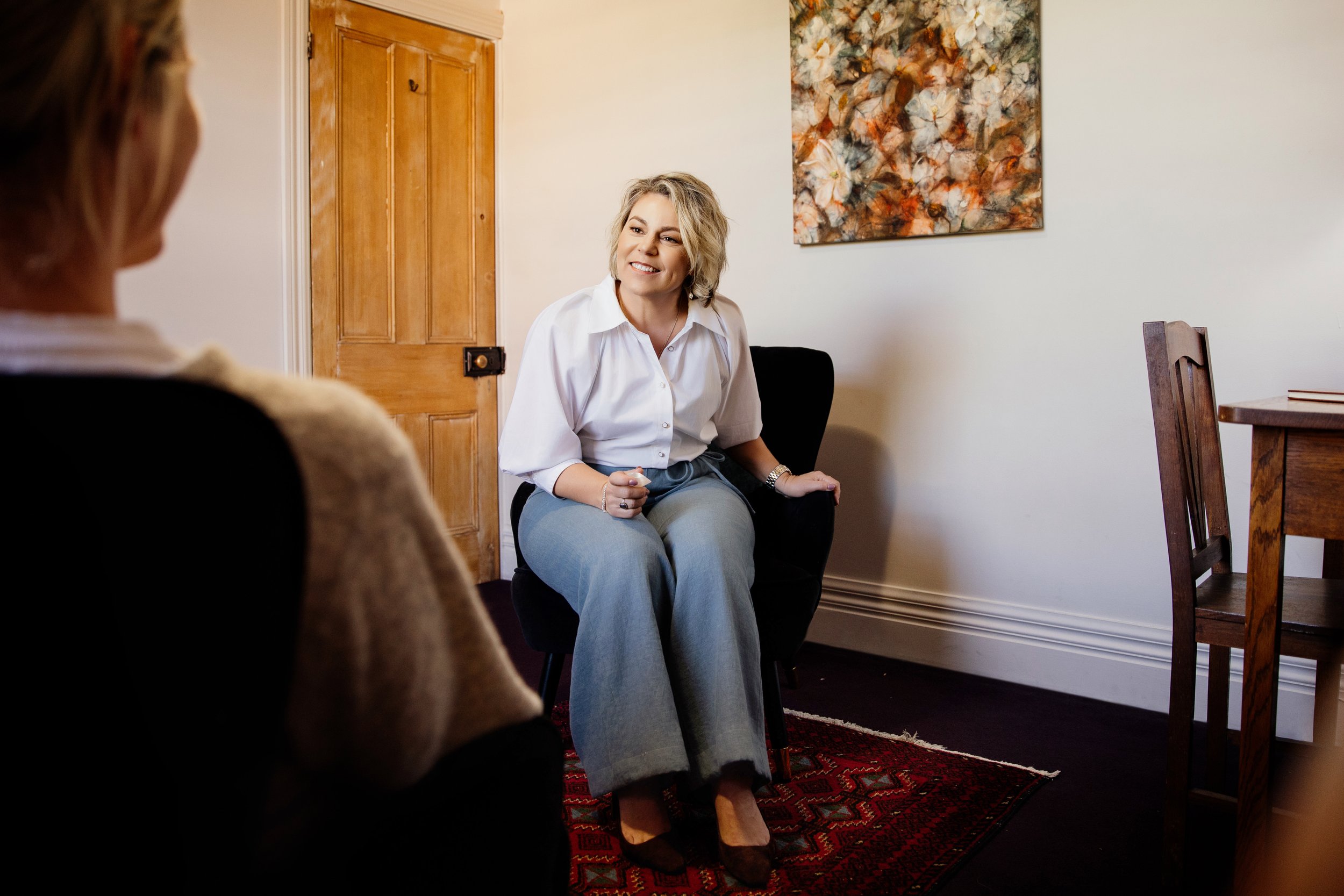 A woman with blonde hair smiling during a conversation in a cozy room, sitting on a black armchair, with a woman whose back is turned visible in the foreground, and a wooden door and colorful abstract art on the wall in the background.