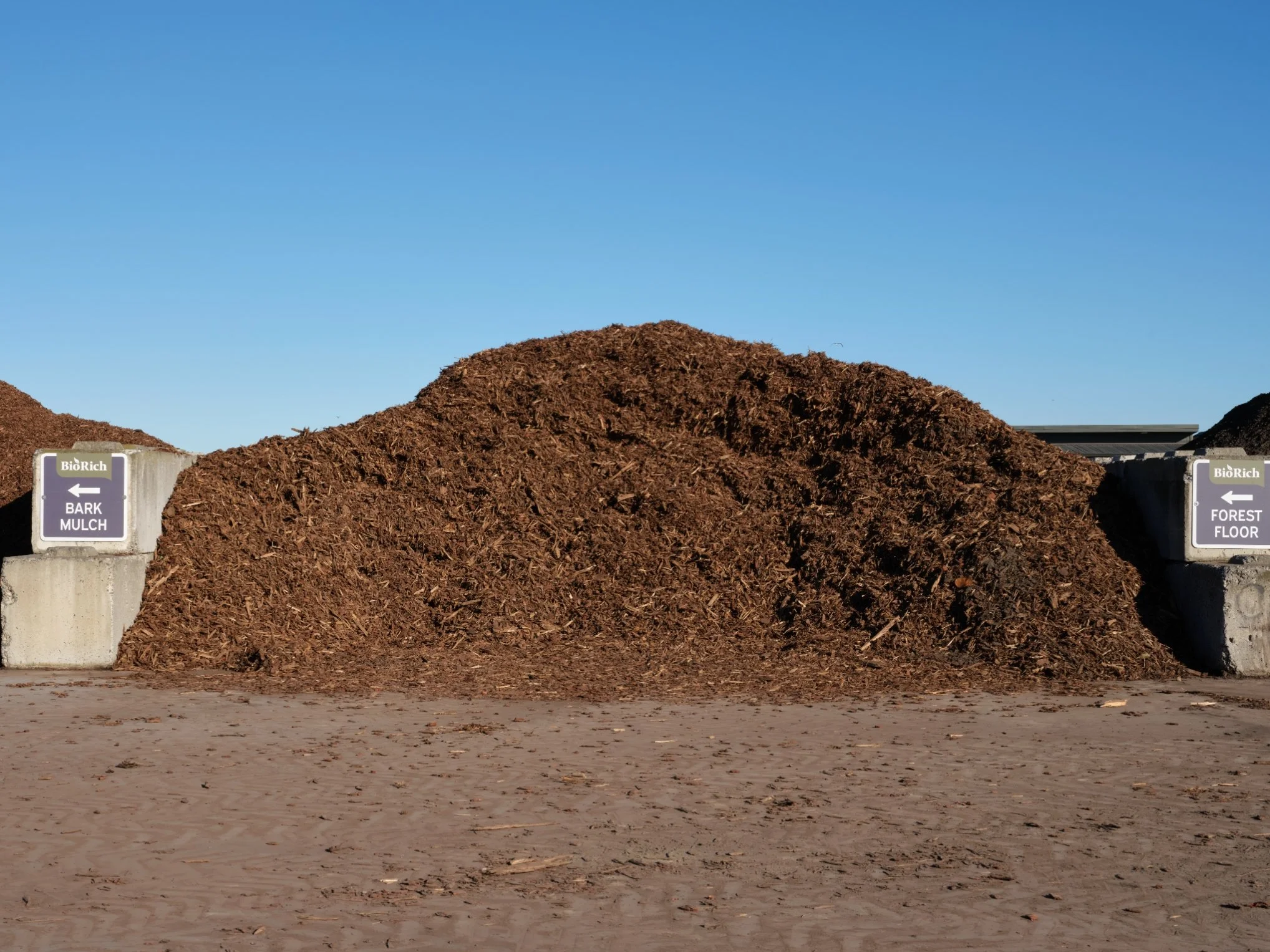 Large pile of mulch in an outdoor area under a clear blue sky with signs indicating directions to bark mulch and forest floor.