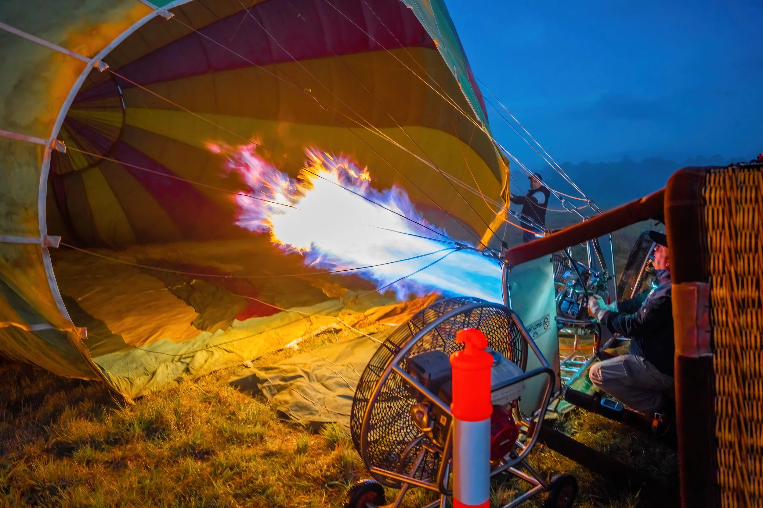 Person inflating a hot air balloon using a torch, with flames visible inside the balloon fabric during dusk.