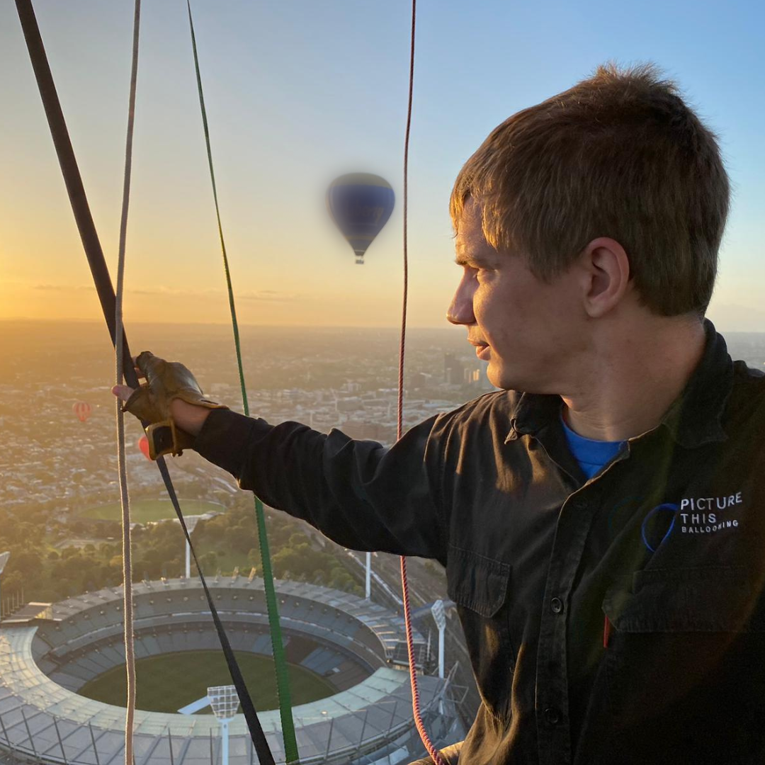 A young man in a black shirt holding a hot air balloon burner at sunset, with a stadium and cityscape below and other hot air balloons in the sky.