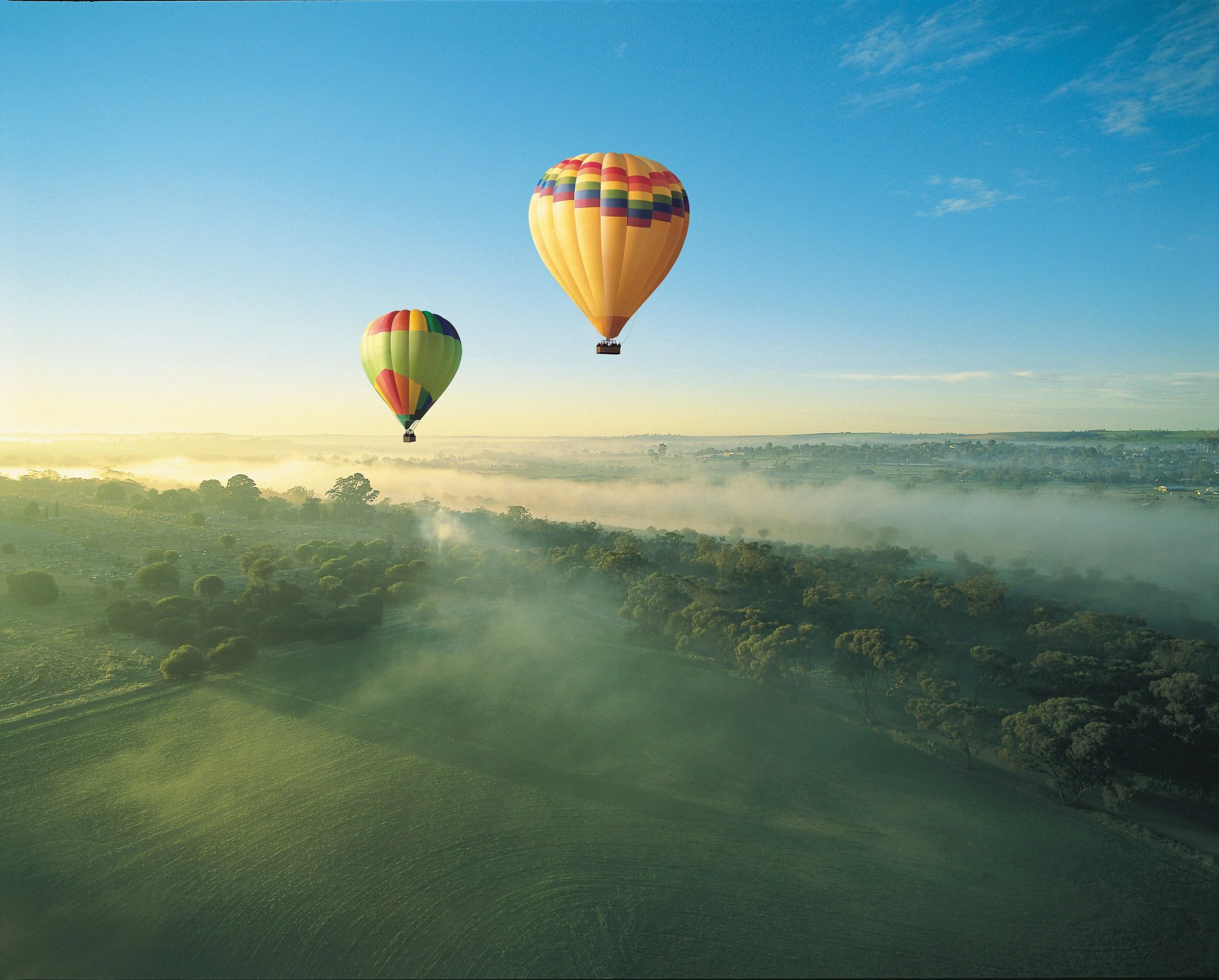 Two hot air balloons flying over a green landscape with trees and fog at sunrise or sunset.
