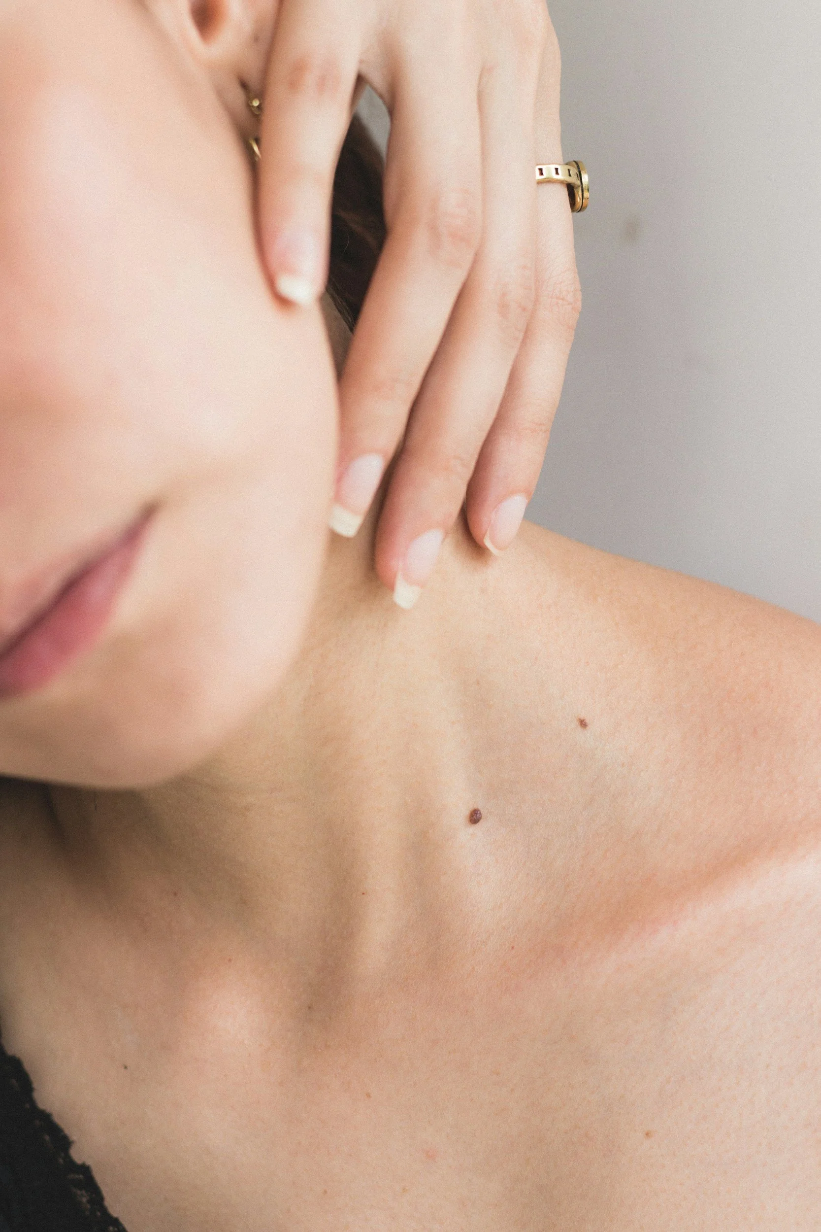 A close-up of a woman's shoulder and neck, with her hand placed on her neck, showing gold rings on her fingers and some small moles on her skin.