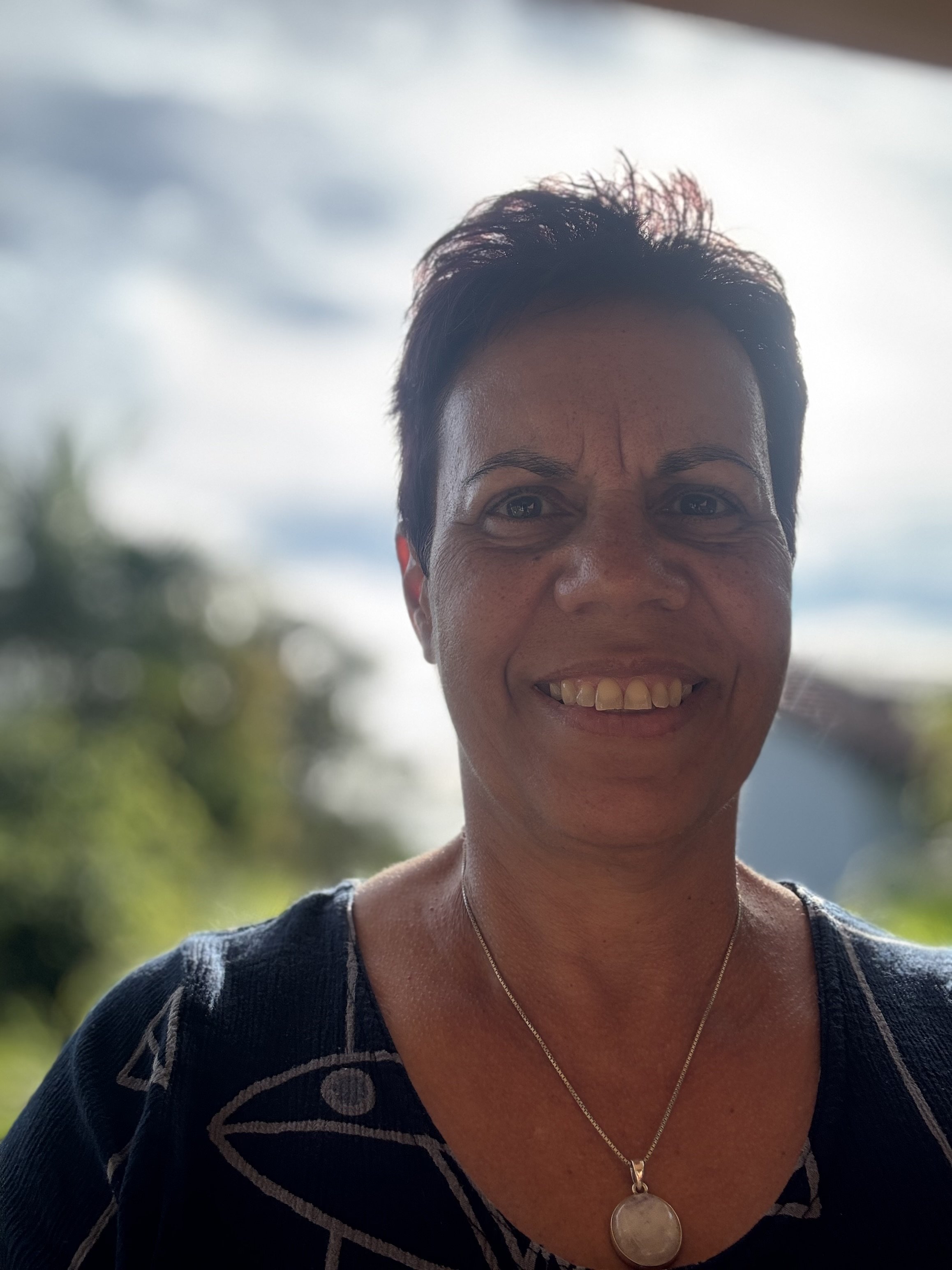 Close-up of a smiling woman with short dark hair, wearing a black patterned top and a silver necklace with a round pendant, outdoors with blurred greenery and cloudy sky in the background.