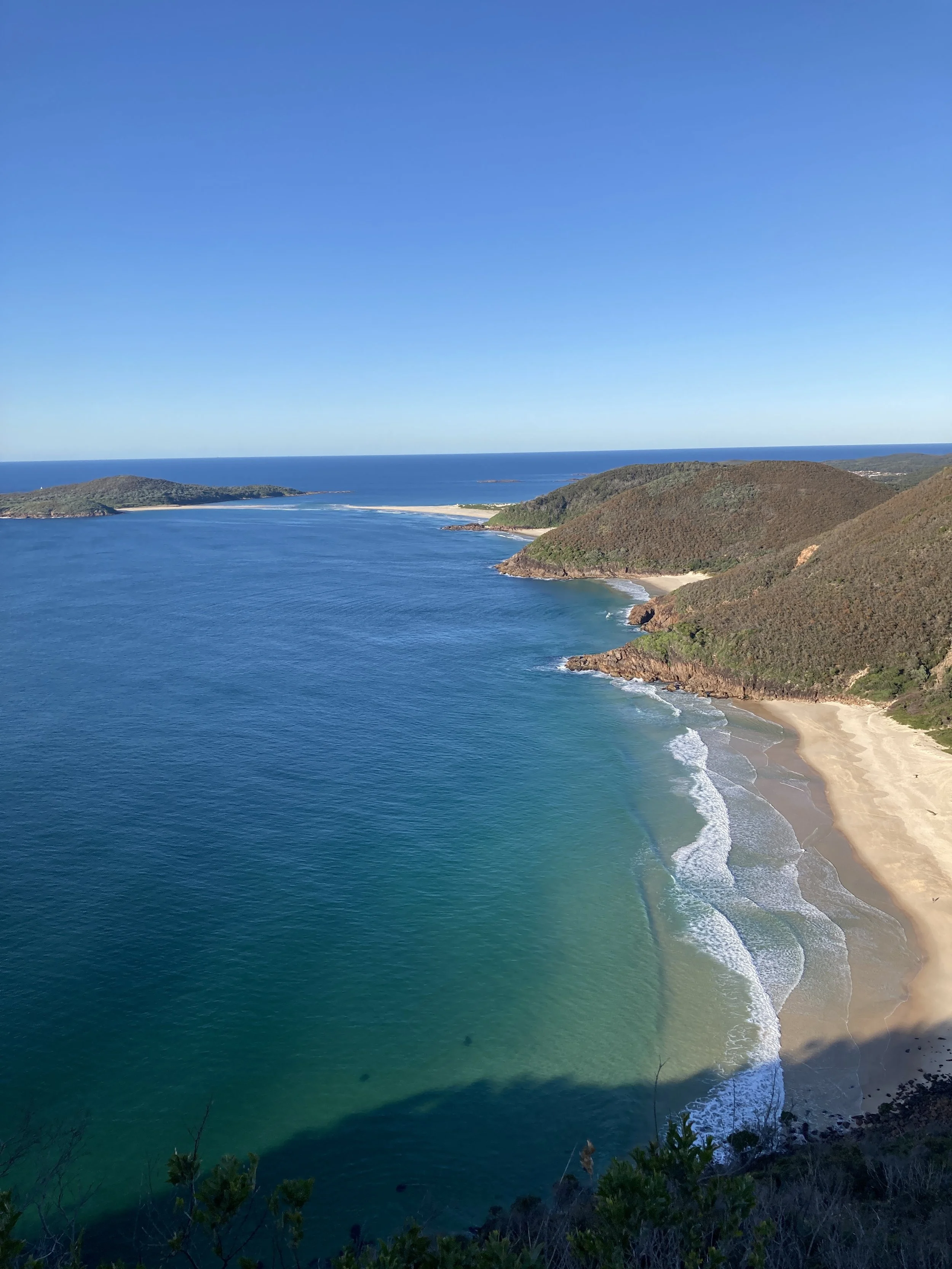 A scenic view of a coastline with blue ocean water, sandy beaches, and green hills under a clear blue sky.