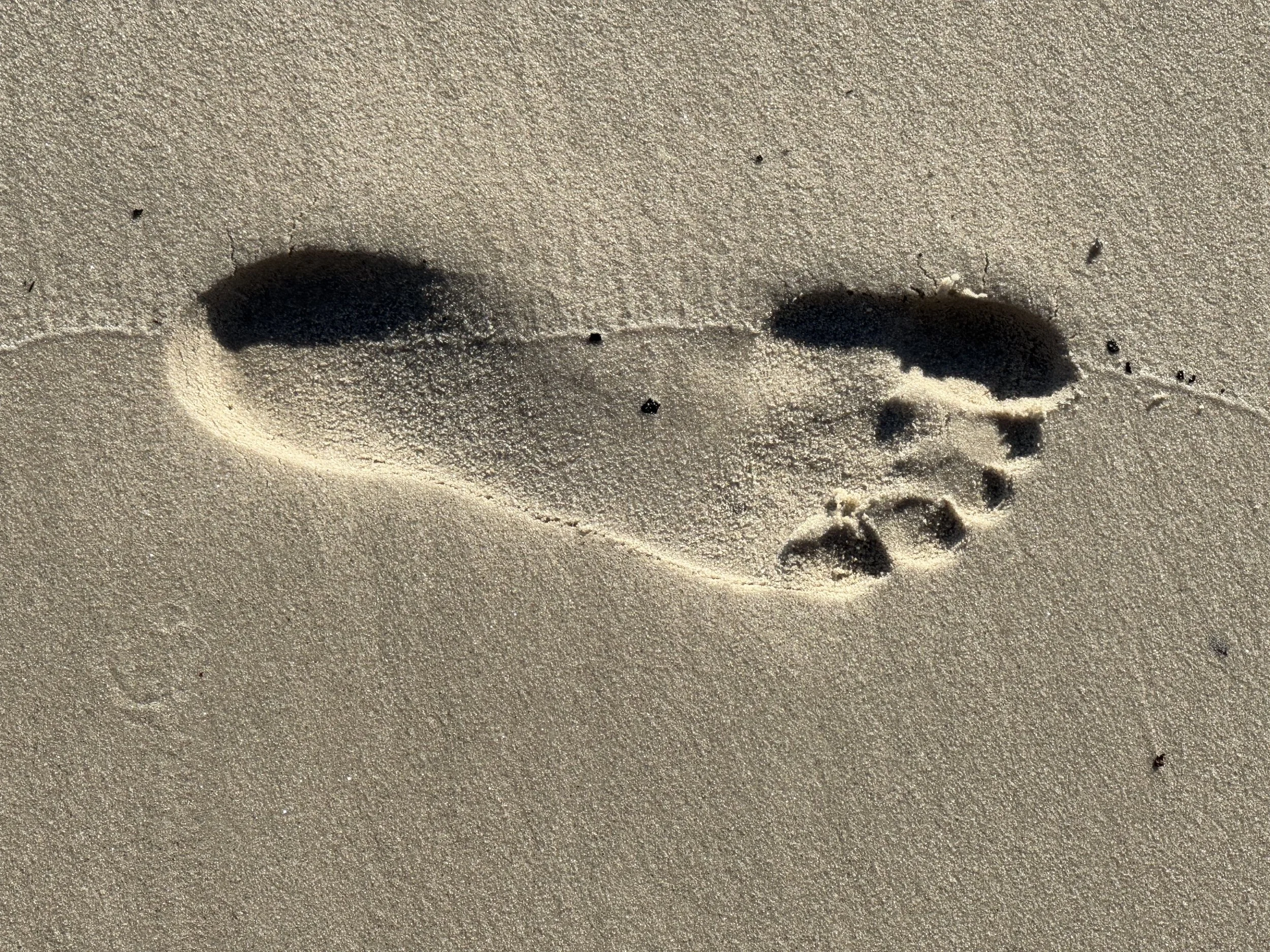 A sand footprint with the toes and heel visible, casting a shadow on the sand.