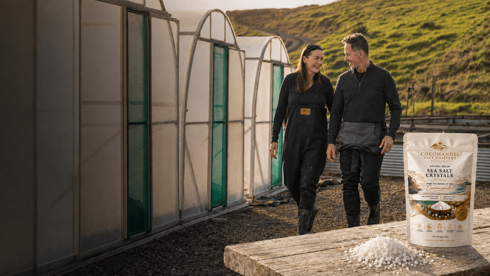 Two people walking outdoors near greenhouses, smiling and enjoying each other's company. A package of sea salt crystals from Coromandel Salt Company and a pile of salt are on a wooden table in the foreground.
