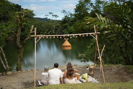 Two people sitting on a blanket by a lake, with a makeshift wooden frame hanging string lights and a basket lamp, surrounded by trees.
