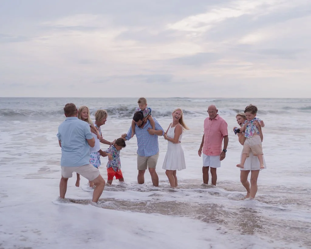 A happy family playing in the ocean waves at the beach, including children, adults, and a baby, during overcast weather.