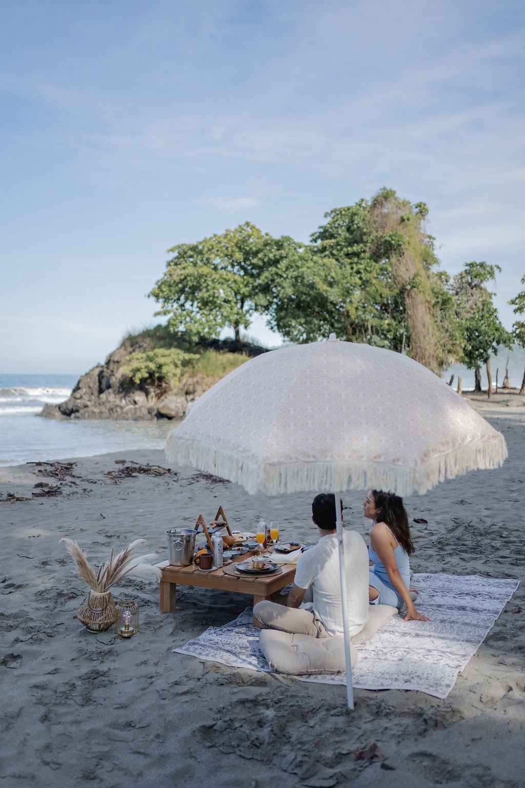 A couple has a picnic on the beach under a large white umbrella. They are sitting on a blanket with a low table set with food, drinks, and utensils. The background features a rocky outcrop, green trees, and a calm ocean under a clear sky.