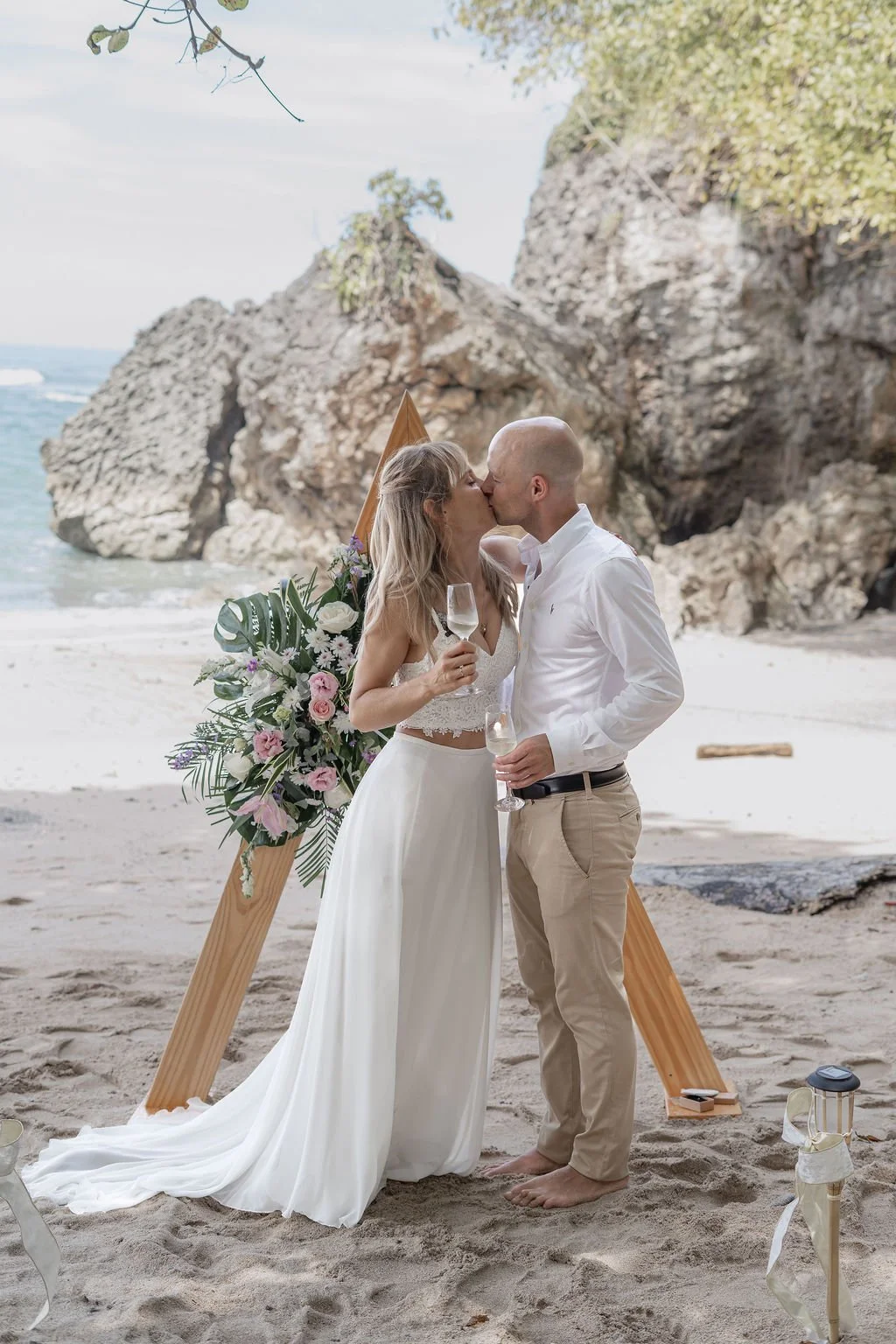 A couple kissing at a beach wedding ceremony with a floral arrangement and a wooden backdrop, ocean and rocks in the background.