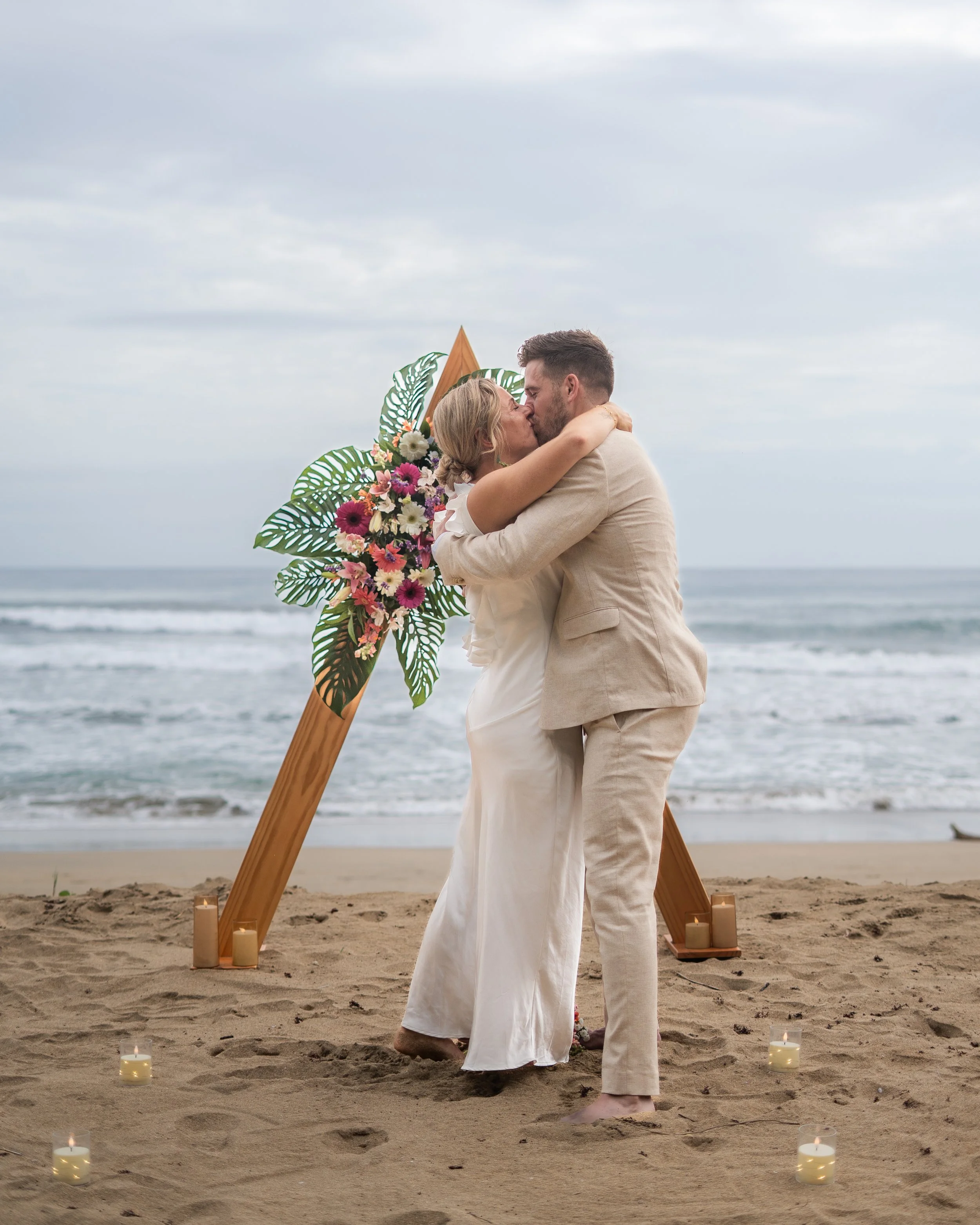 A wedding ceremony taking place outdoors among lush green tropical plants with a man in a white shirt and a woman in a white wedding dress, holding a book.