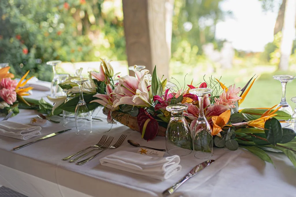 Table with a floral centerpiece, wine glasses upside down, white napkins, and silverware set for a formal event, outdoors with trees in the background.