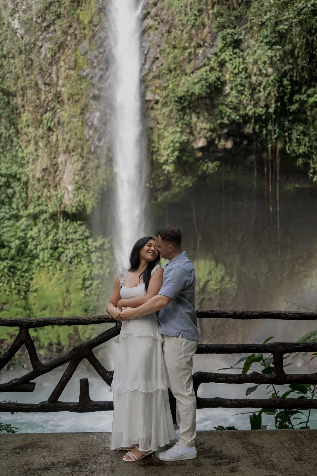 A couple embracing in front of a waterfall with lush green foliage and a wooden railing.