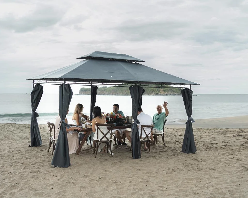 Group of people having a toast at an outdoor beachside dining table under a canopy, with a floral centerpiece, glasses of beer, and a beach with greenery in the background.
