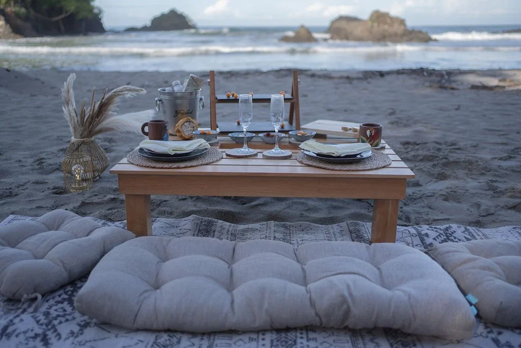 A beach picnic setup with a low wooden table, placed on a patterned blanket and surrounded by cushions, set with plates, glasses, mugs, and a bucket containing drinks, with the ocean and rocks in the background.