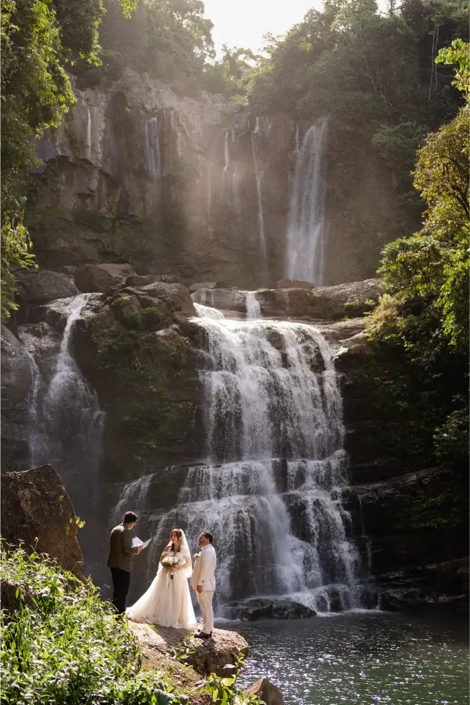 Two people sitting on a blanket by a lake, with a makeshift wooden frame hanging string lights and a basket lamp, surrounded by trees.