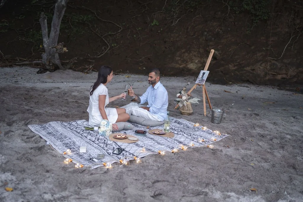 A couple having a romantic picnic on a blanket with food and drinks on a beach at sunset, surrounded by decorative lights and flowers organised by Macaw Experiences in Costa Rica