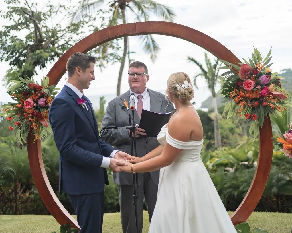 A couple getting married outdoors, holding hands, with an officiant standing behind them, under a circular floral arch with tropical flowers, in a lush green area with palm trees. In Costa Rica organised by Macaw Experiences