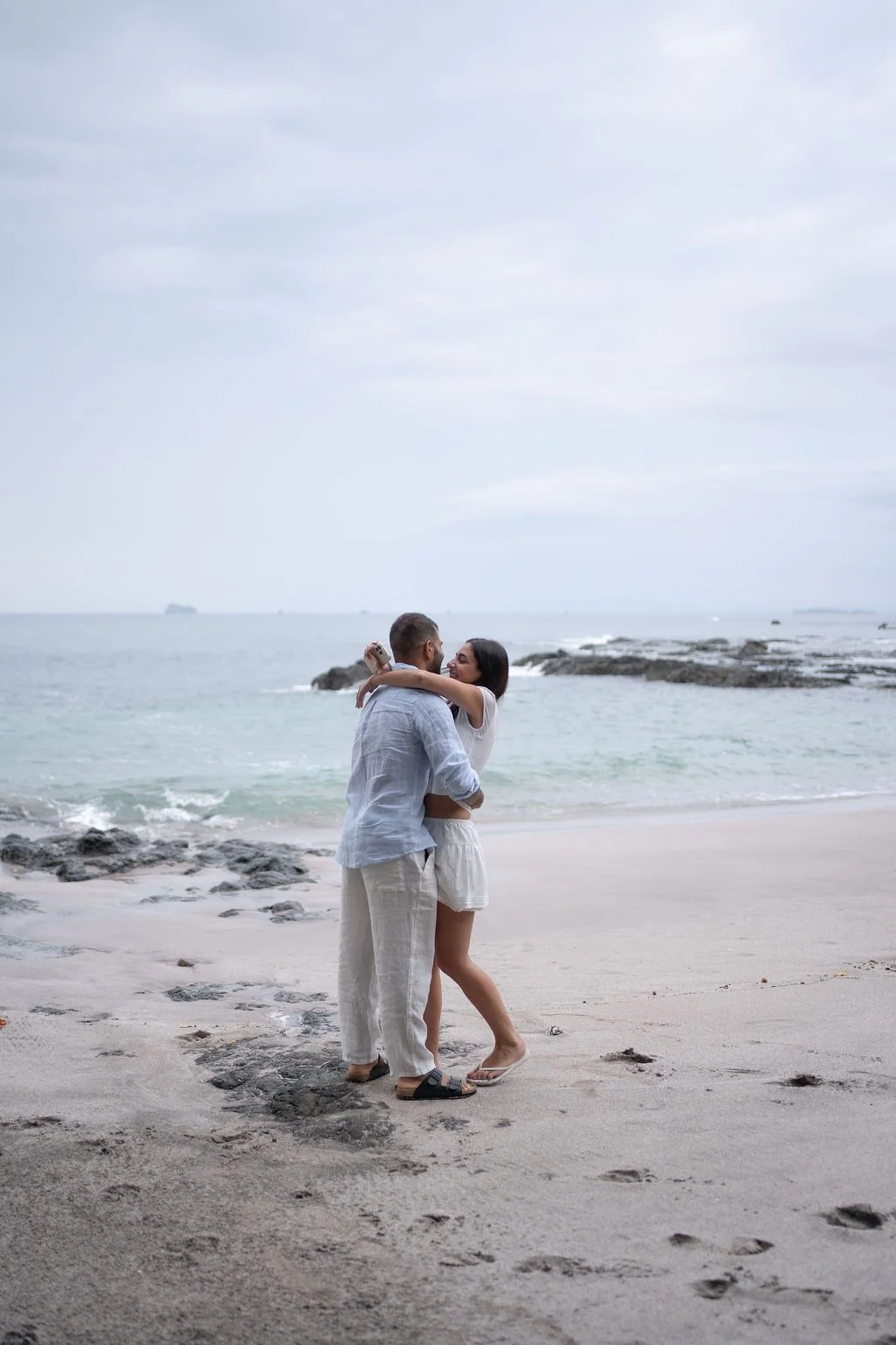A couple embraces on a beach, with the ocean and rocky shoreline in the background, under a cloudy sky.