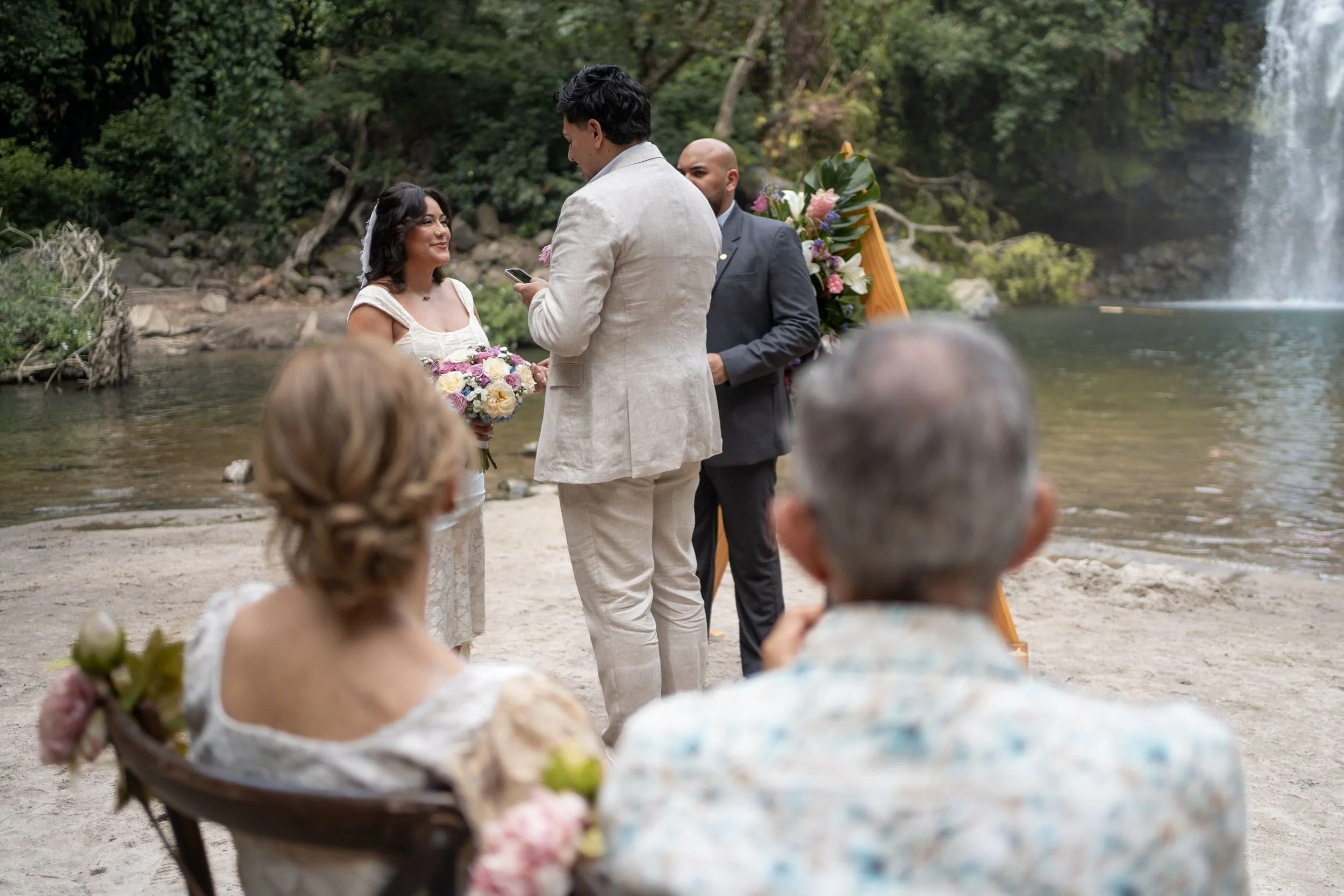 A couple getting married on a beach, with a person officiating and a decorative wooden wedding arch decorated with flowers behind them in Costa Rica