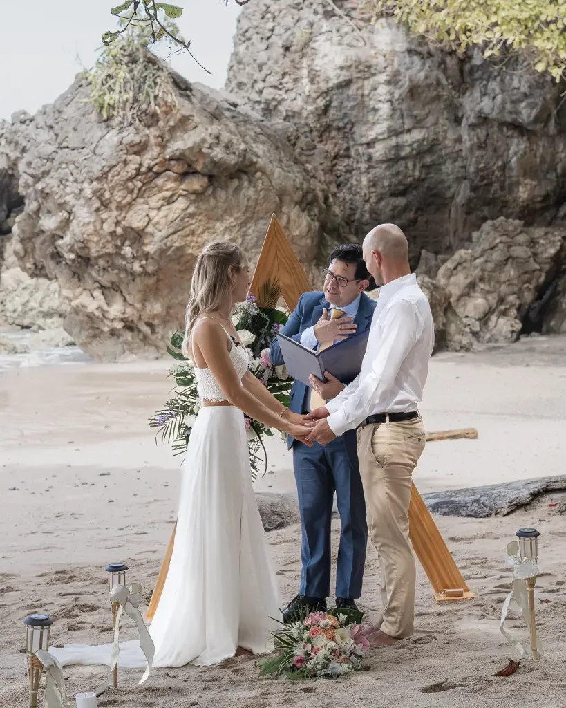 A couple getting married on a beach, with a person officiating and a decorative wooden wedding arch decorated with flowers behind them in Costa Rica