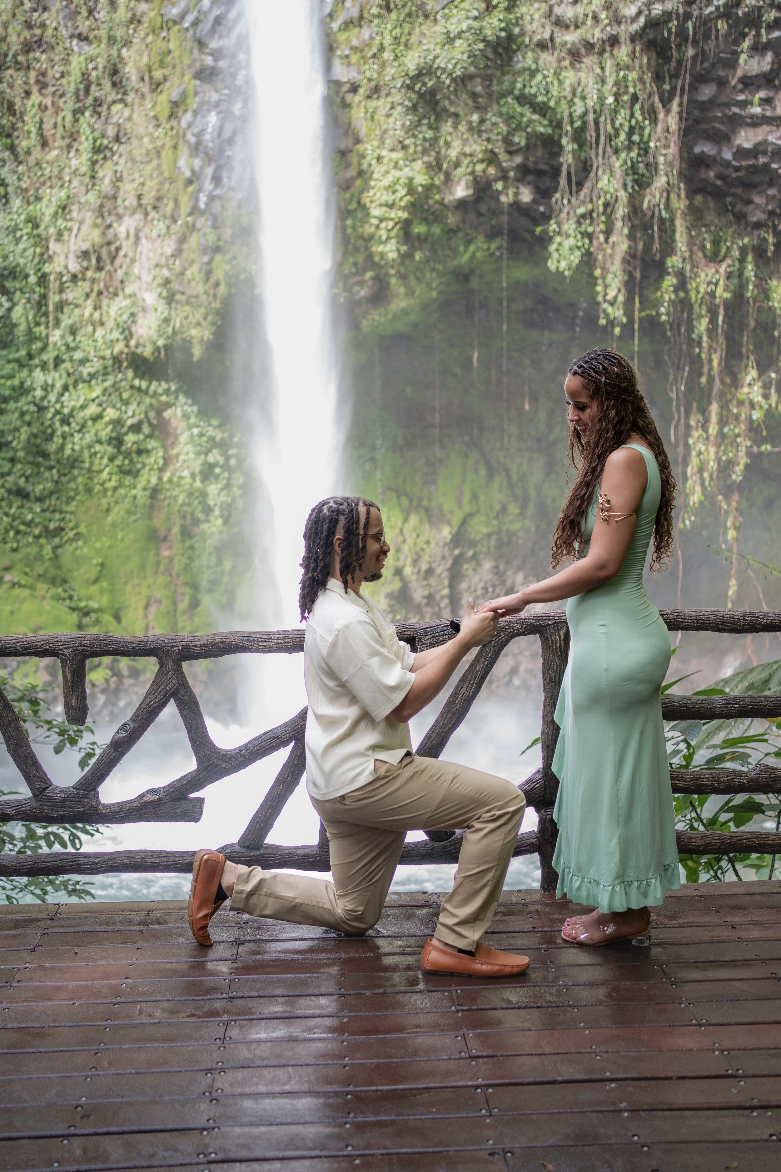 A man proposing to a woman on a wooden platform in front of a waterfall surrounded by lush greenery.