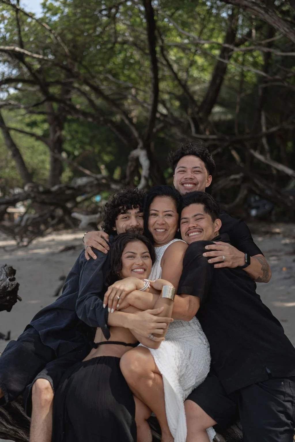Group of five people hugging and smiling on a beach with a large fallen tree and green trees in the background.