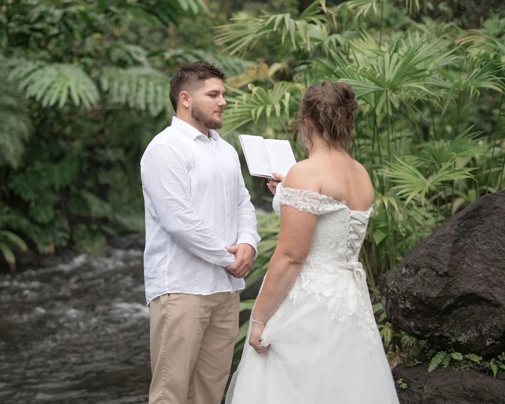 A wedding ceremony taking place outdoors among lush green tropical plants with a man in a white shirt and a woman in a white wedding dress, holding a book.