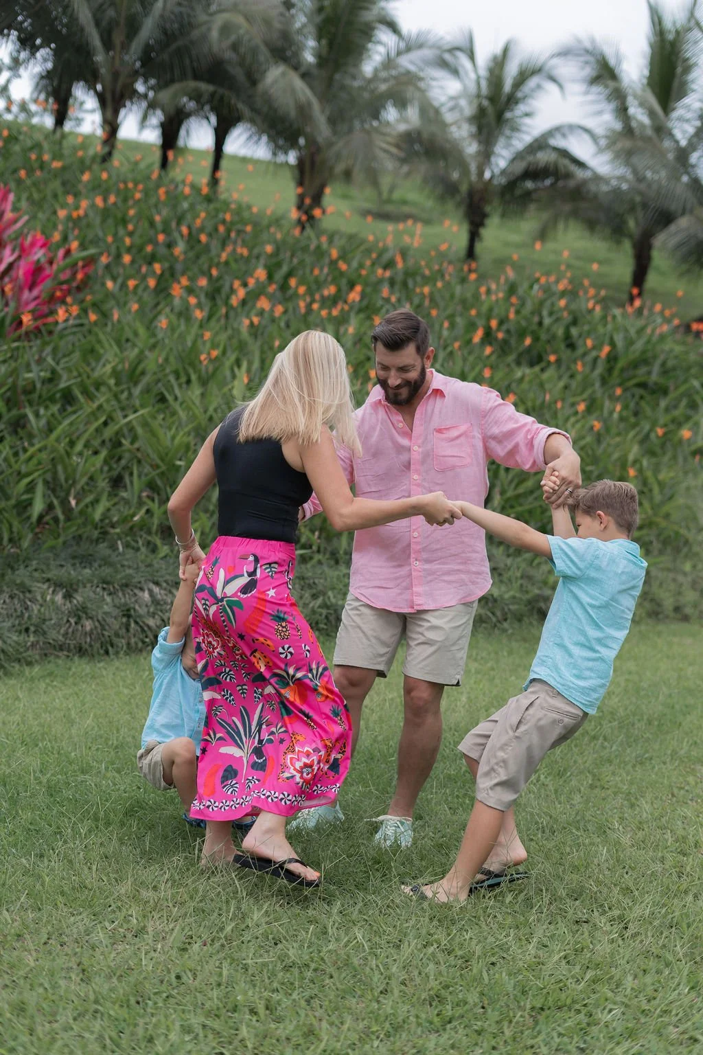 A family of four playing and holding hands in a grassy field with tropical plants and palm trees in the background.