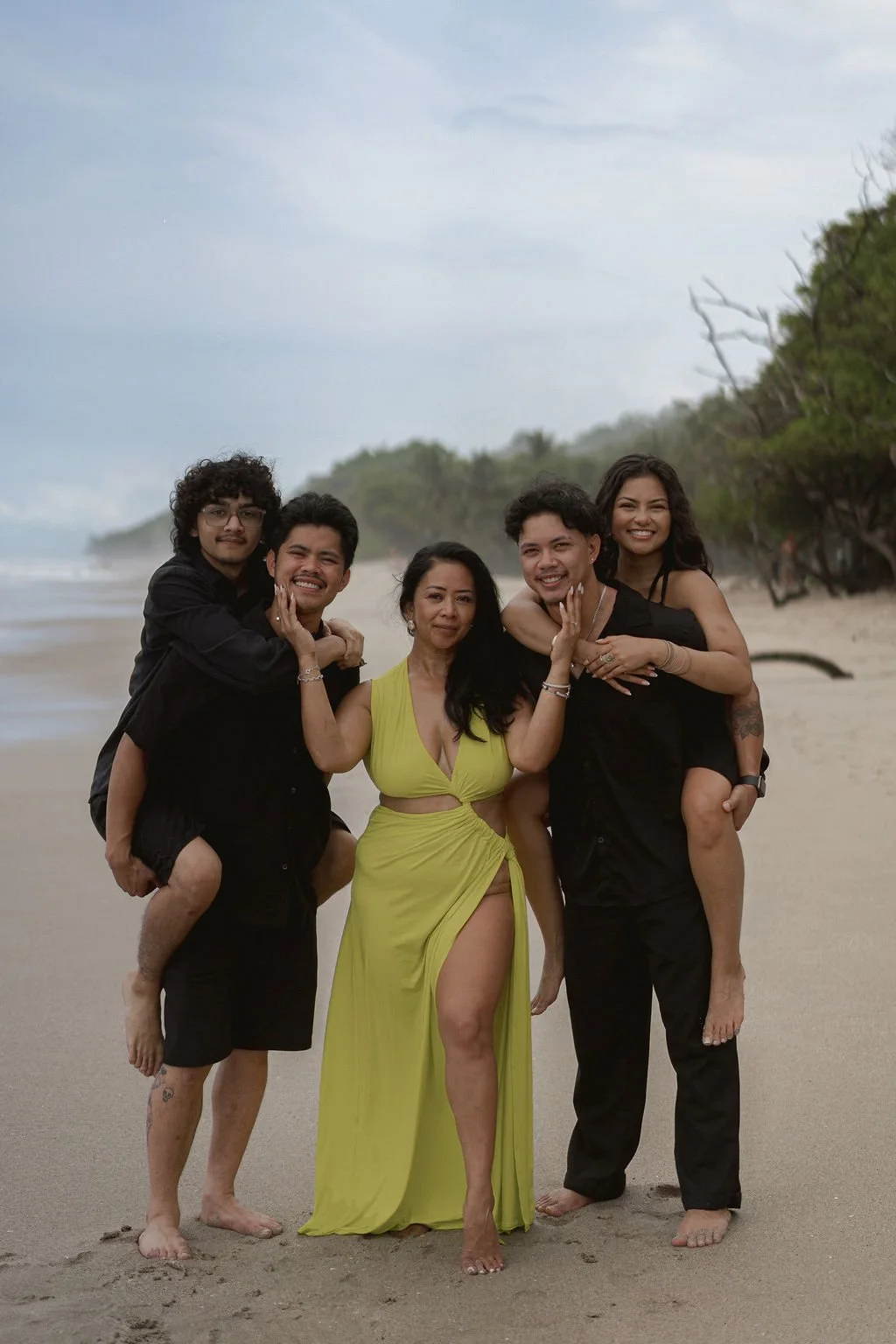Group of five friends at the beach, smiling and posing together, with ocean and trees in the background