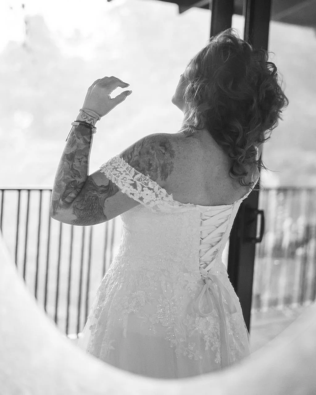 A woman with tattoos on her arm wearing a lace wedding dress, seen from behind, adjusting her hair.