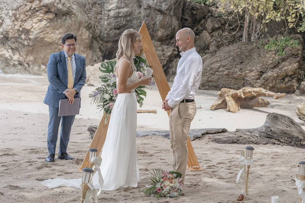 A couple getting married on a beach, with a person officiating and a decorative wooden wedding arch decorated with flowers behind them in Costa Rica