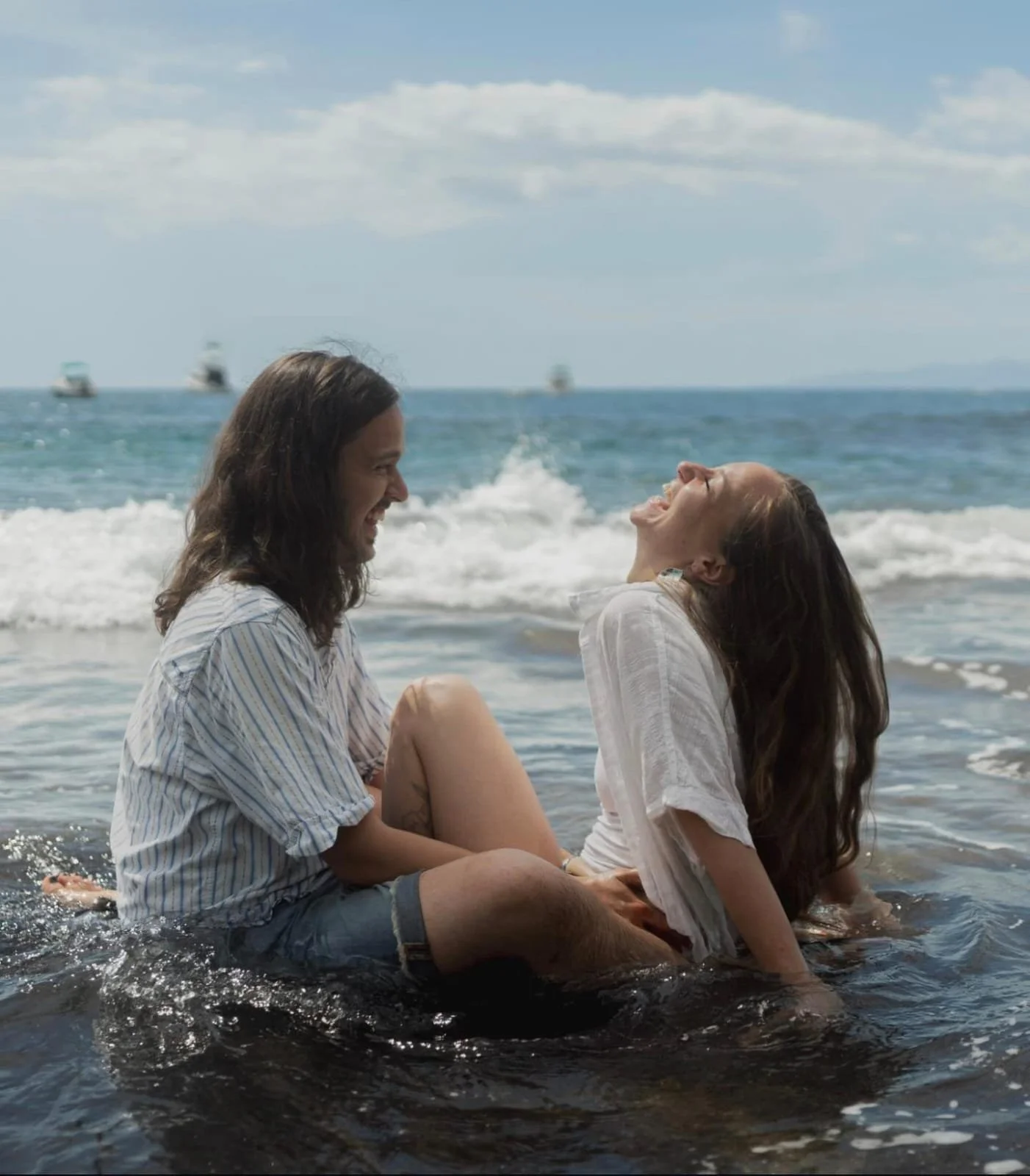 A couple smiling and laughing while sitting in shallow ocean water at the beach, with boats on the horizon and partly cloudy sky.