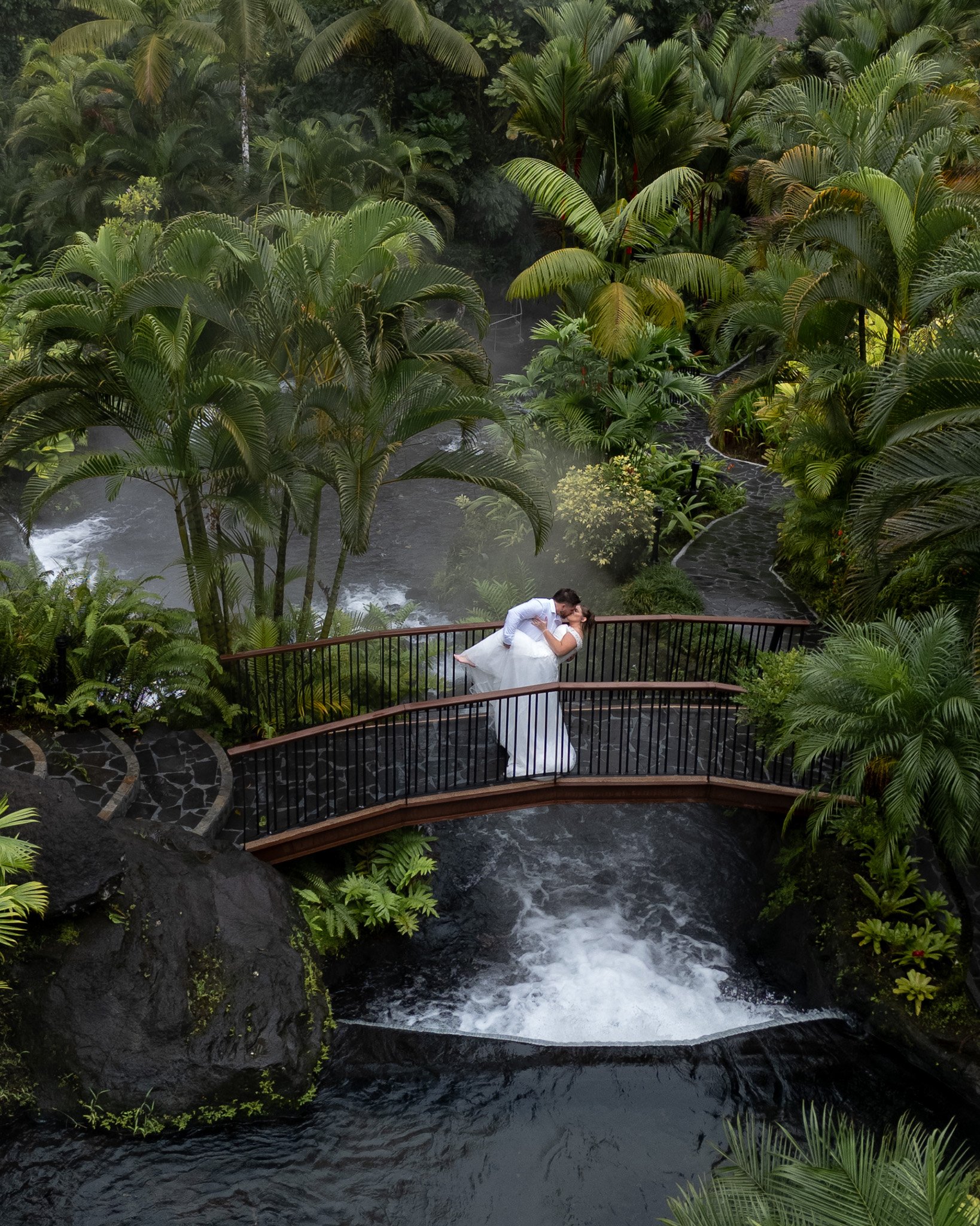 A wedding ceremony taking place outdoors among lush green tropical plants with a man in a white shirt and a woman in a white wedding dress, holding a book.