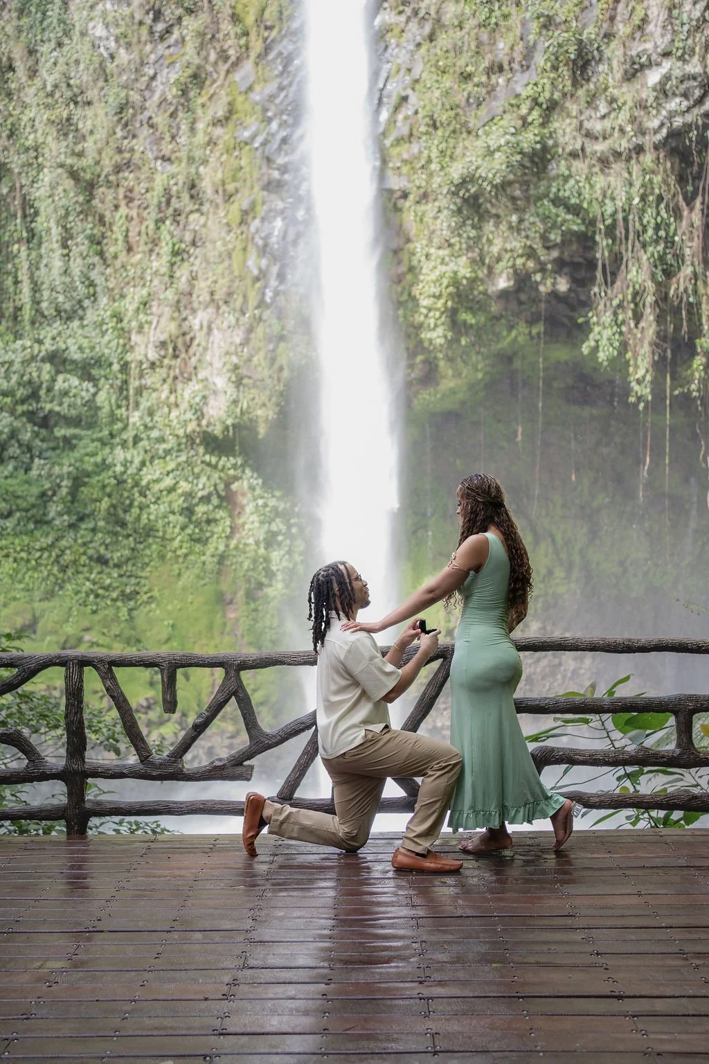 A man proposes marriage to a woman on a wooden platform in front of a waterfall surrounded by lush greenery in Costa Rica