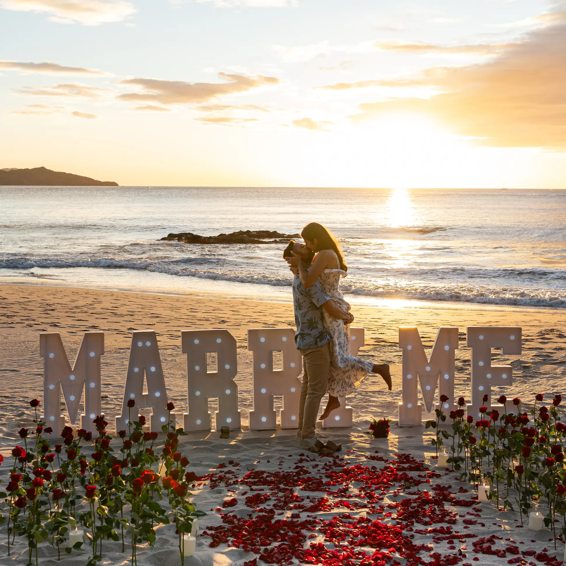 A couple has a picnic on the beach under a large white umbrella. They are sitting on a blanket with a low table set with food, drinks, and utensils. The background features a rocky outcrop, green trees, and a calm ocean under a clear sky.