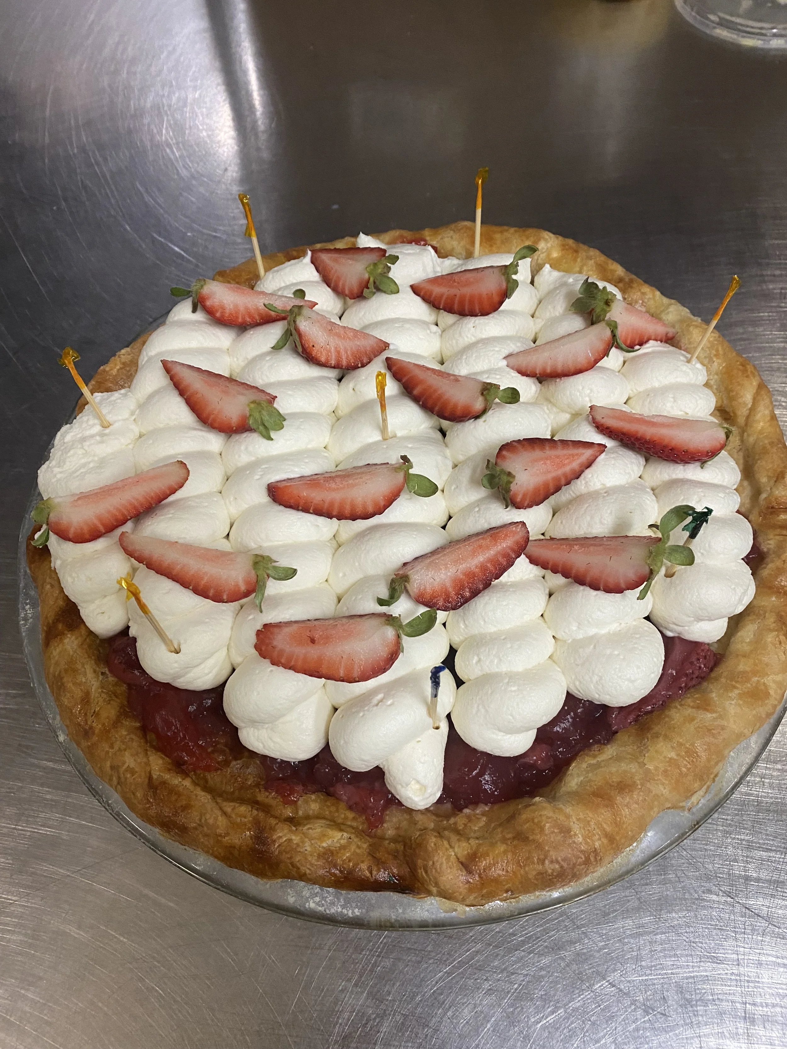 Strawberry tart with a golden crust, topped with whipped cream and fresh sliced strawberries, garnished with small green leaves, placed on a glass plate.