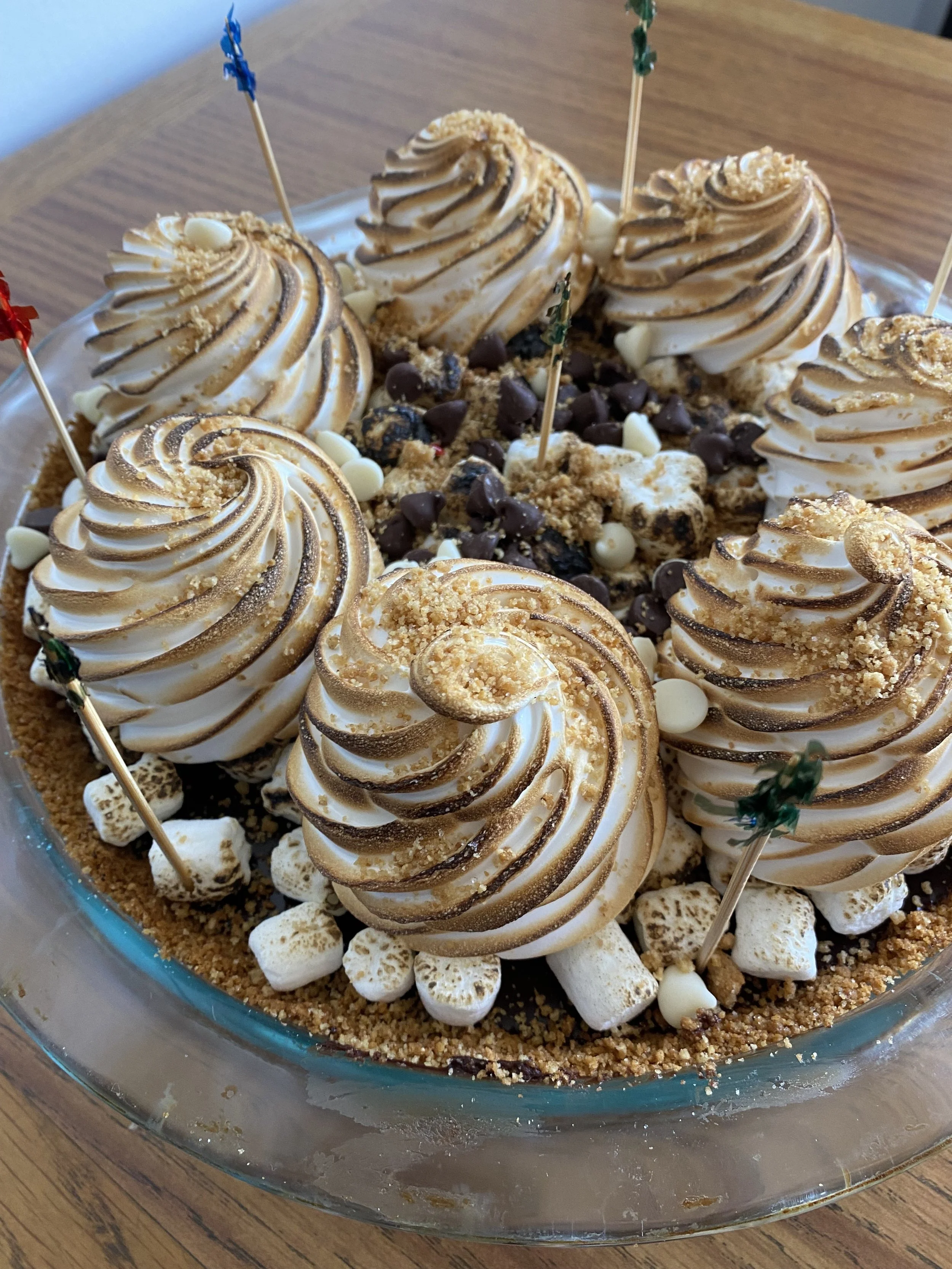 A dessert cake with swirled roasted marshmallows, chocolate chips, cookie crumbs, and white chocolate chips, decorated with colorful toothpick flags.