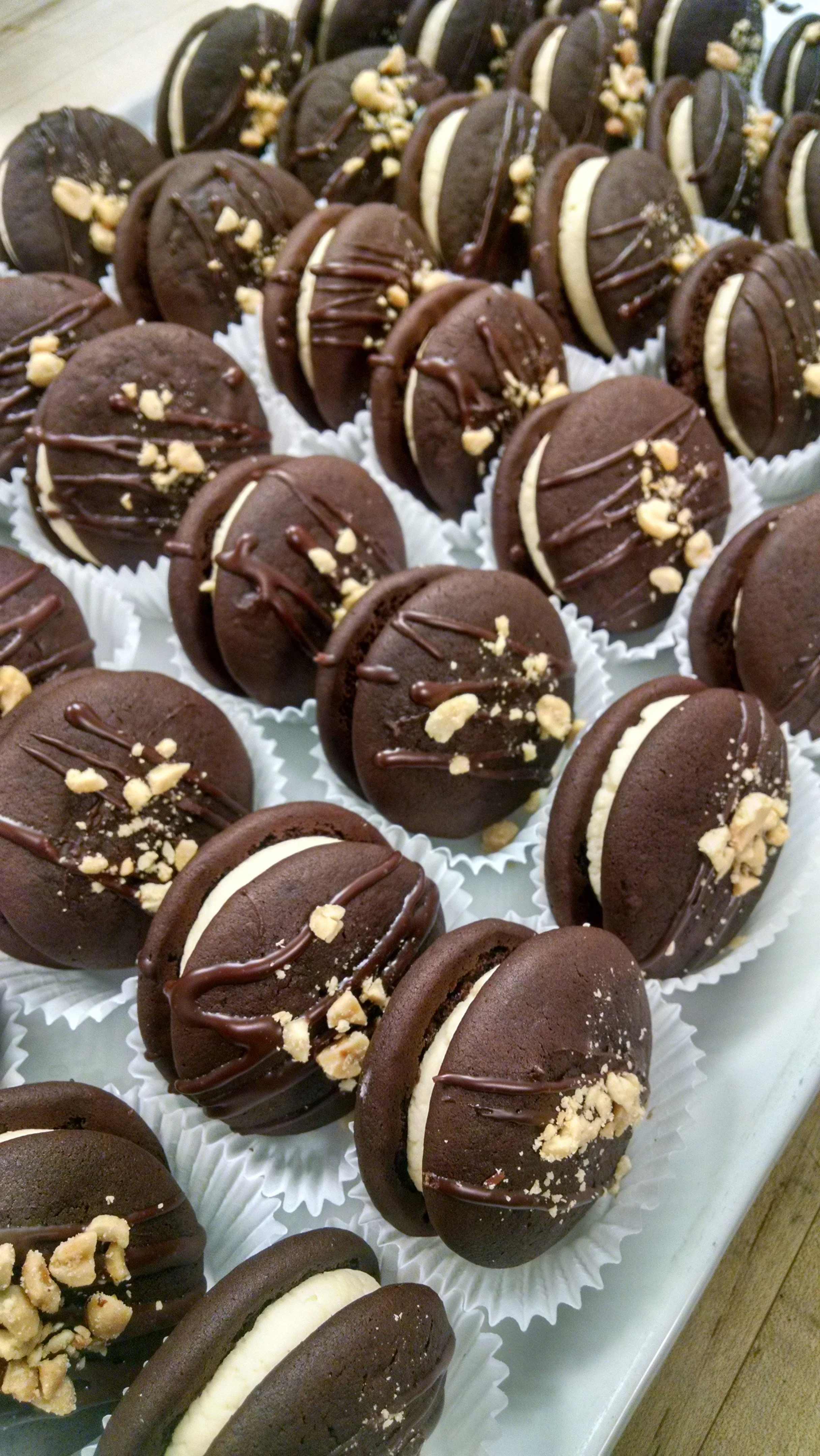 Row of chocolate cookies filled with cream, topped with crushed nuts and drizzled with chocolate.