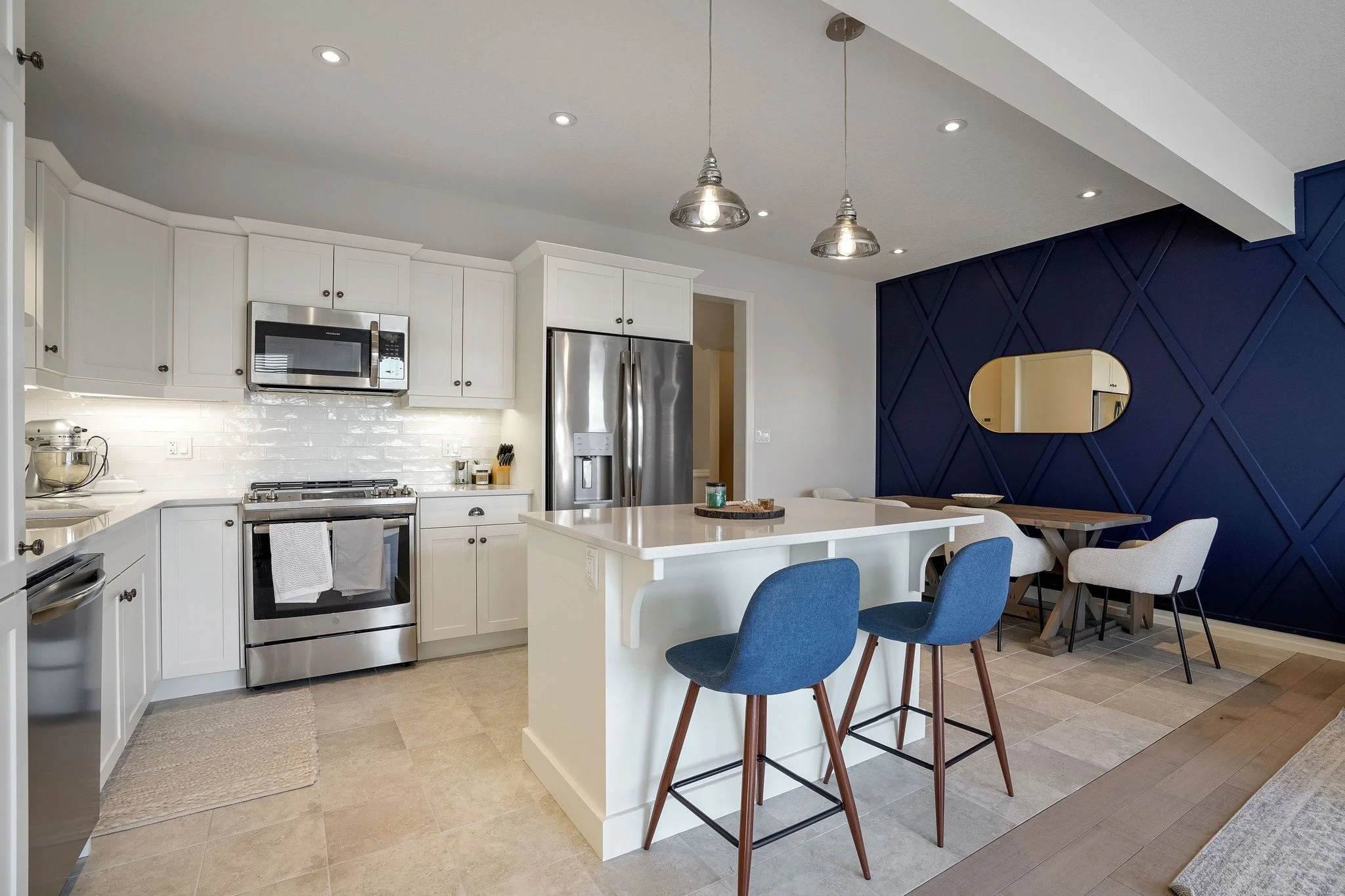 Modern kitchen with white cabinets, stainless steel appliances, a navy accent wall, and a kitchen island with blue and beige chairs.