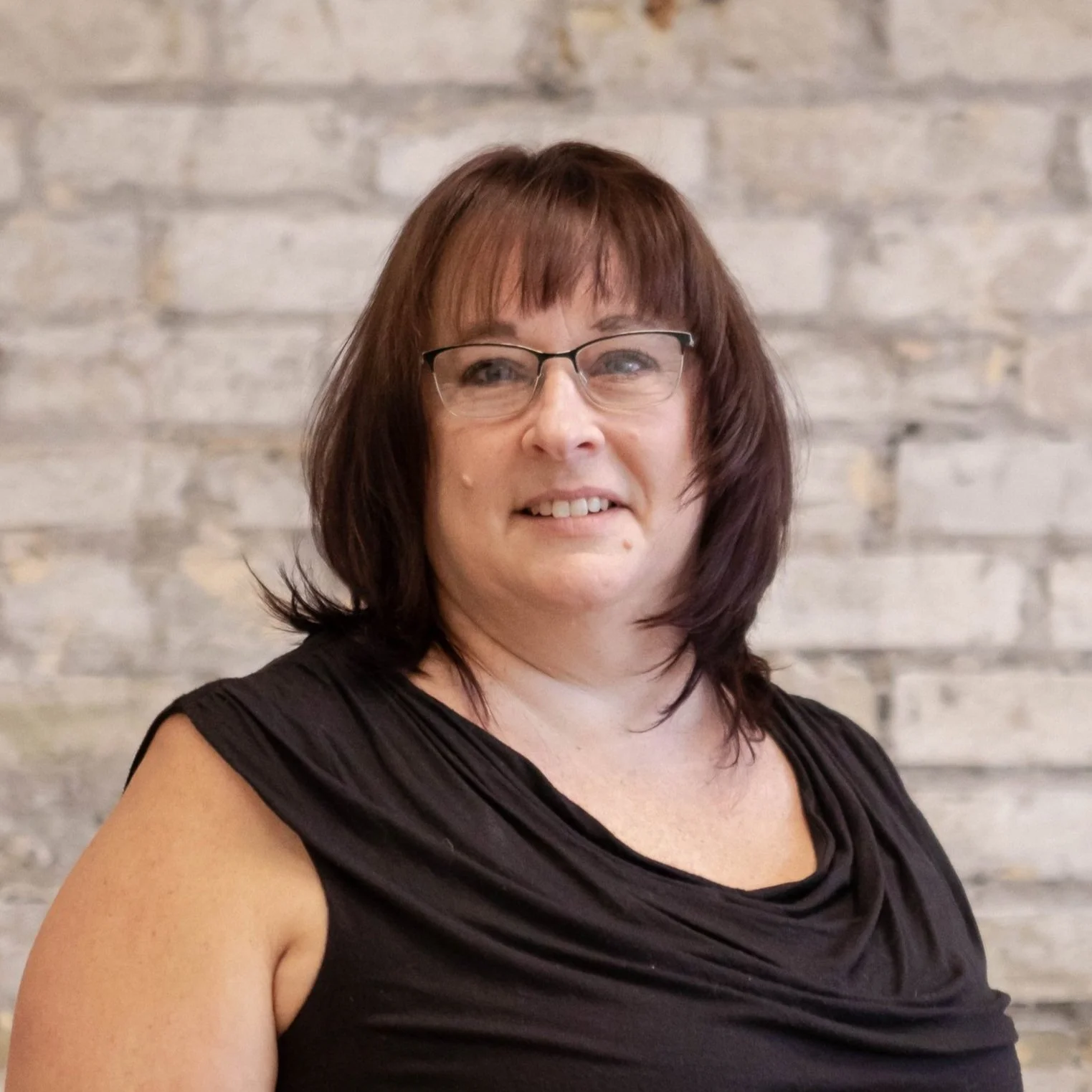 A woman with shoulder-length brown hair and glasses, wearing a black top, smiling in front of a brick wall.