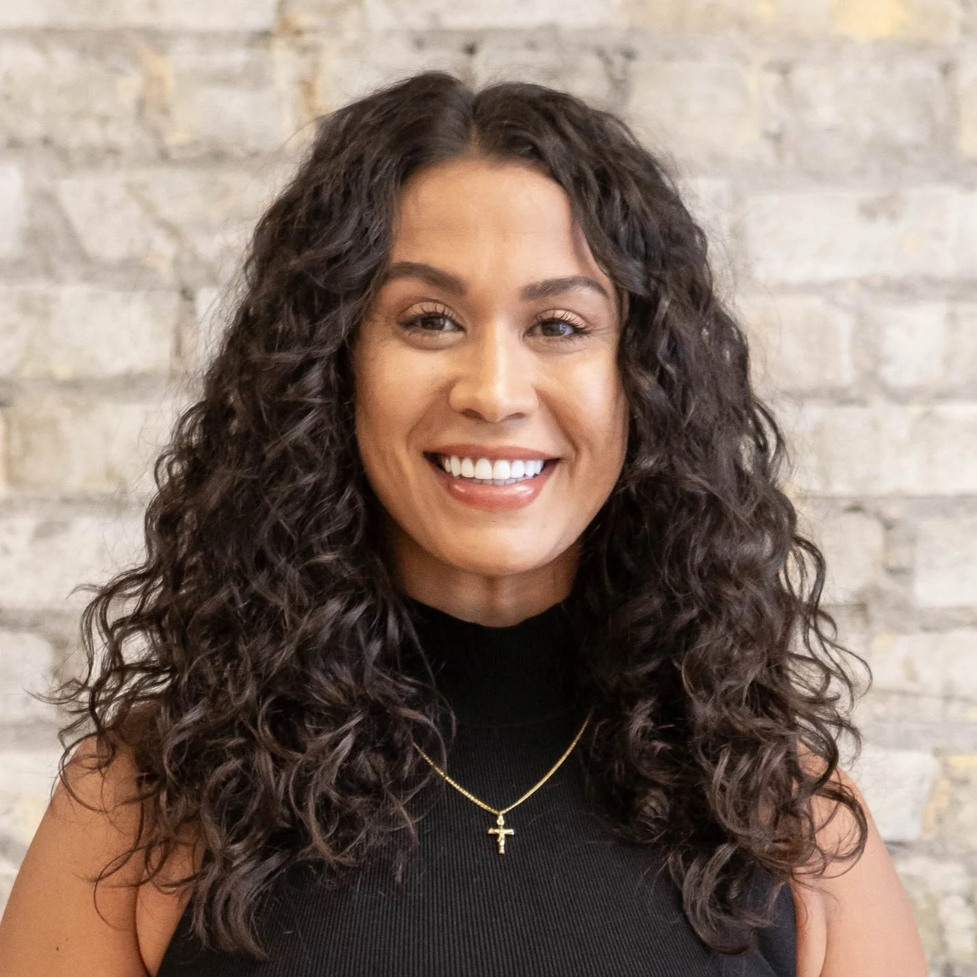 A woman with long, curly dark hair, smiling, wearing a black sleeveless top and a gold necklace with a cross pendant. Background is a light-colored brick wall.