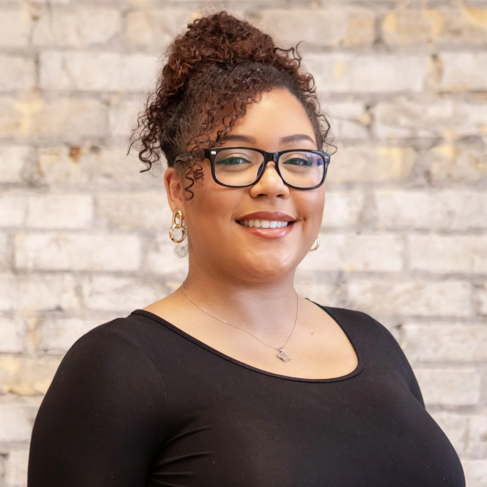 A woman with curly hair in a bun, wearing glasses, earrings, a necklace, and a black top, smiling in front of a brick wall.