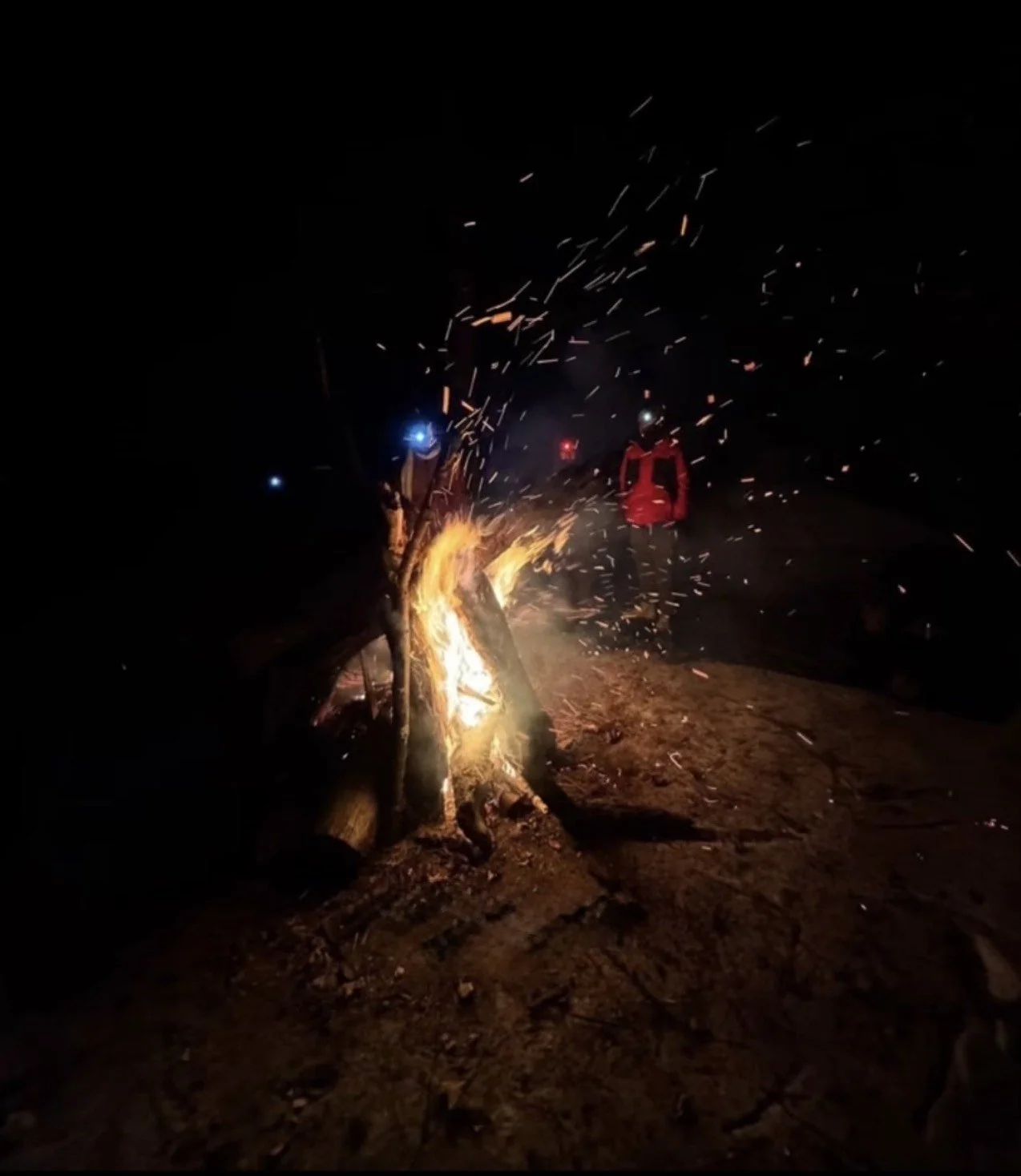 Two people standing near a bonfire at night, with sparks flying in the air and wearing headlamps.